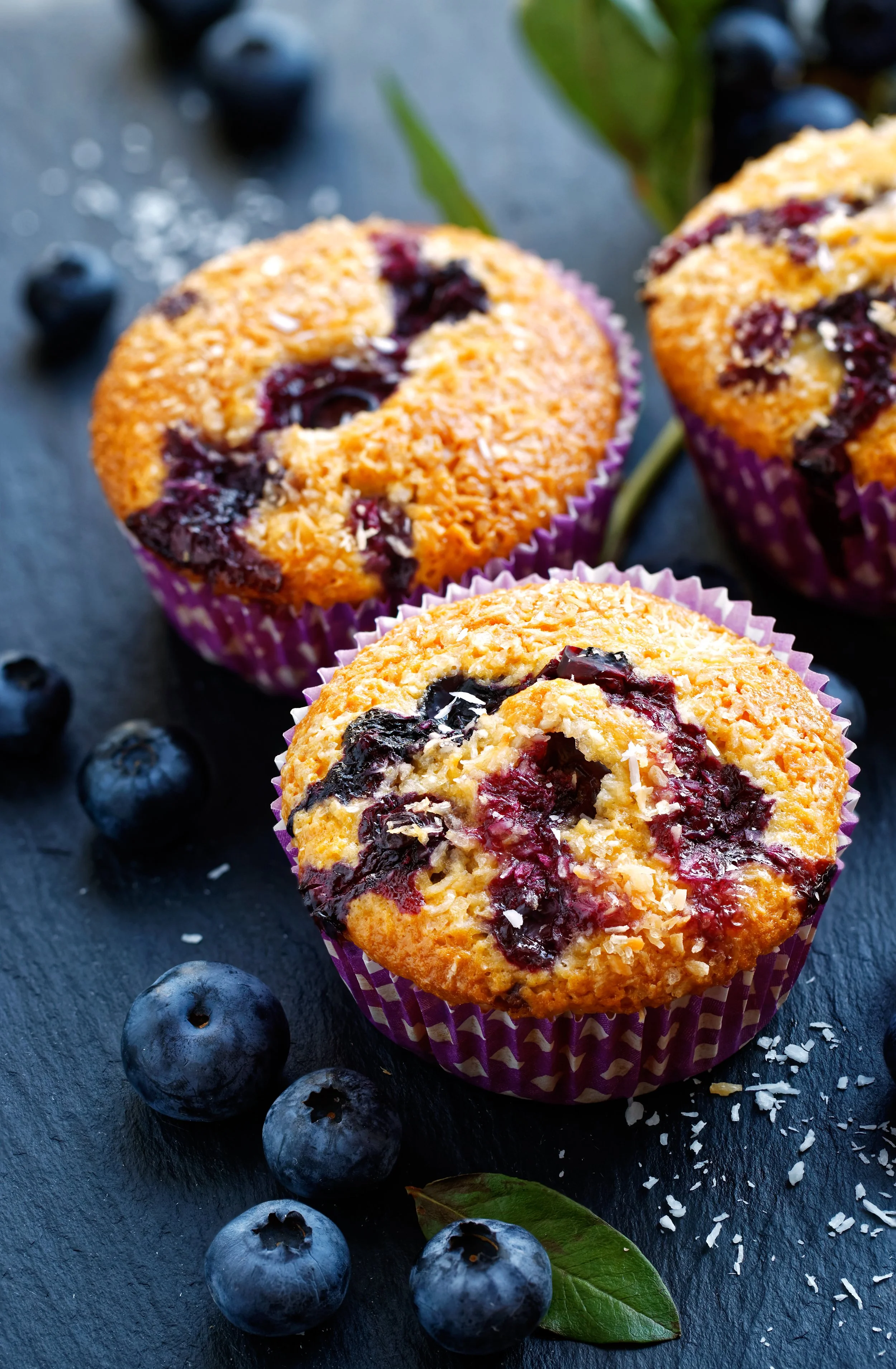 Three blueberry muffins in purple cupcake liners on a black slate surface, with fresh blueberries and a green leaf scattered around.