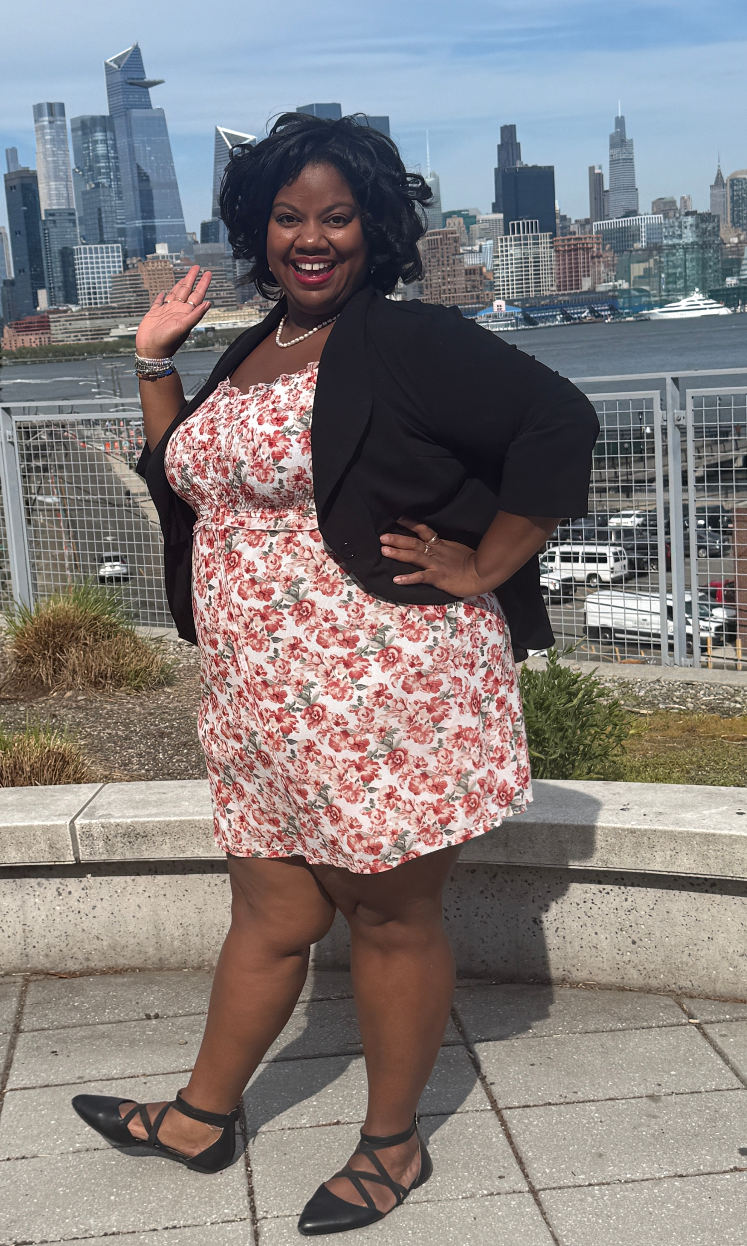 Lissette in a with dress with red roses and a black blazer smiling and waving against a city backdrop