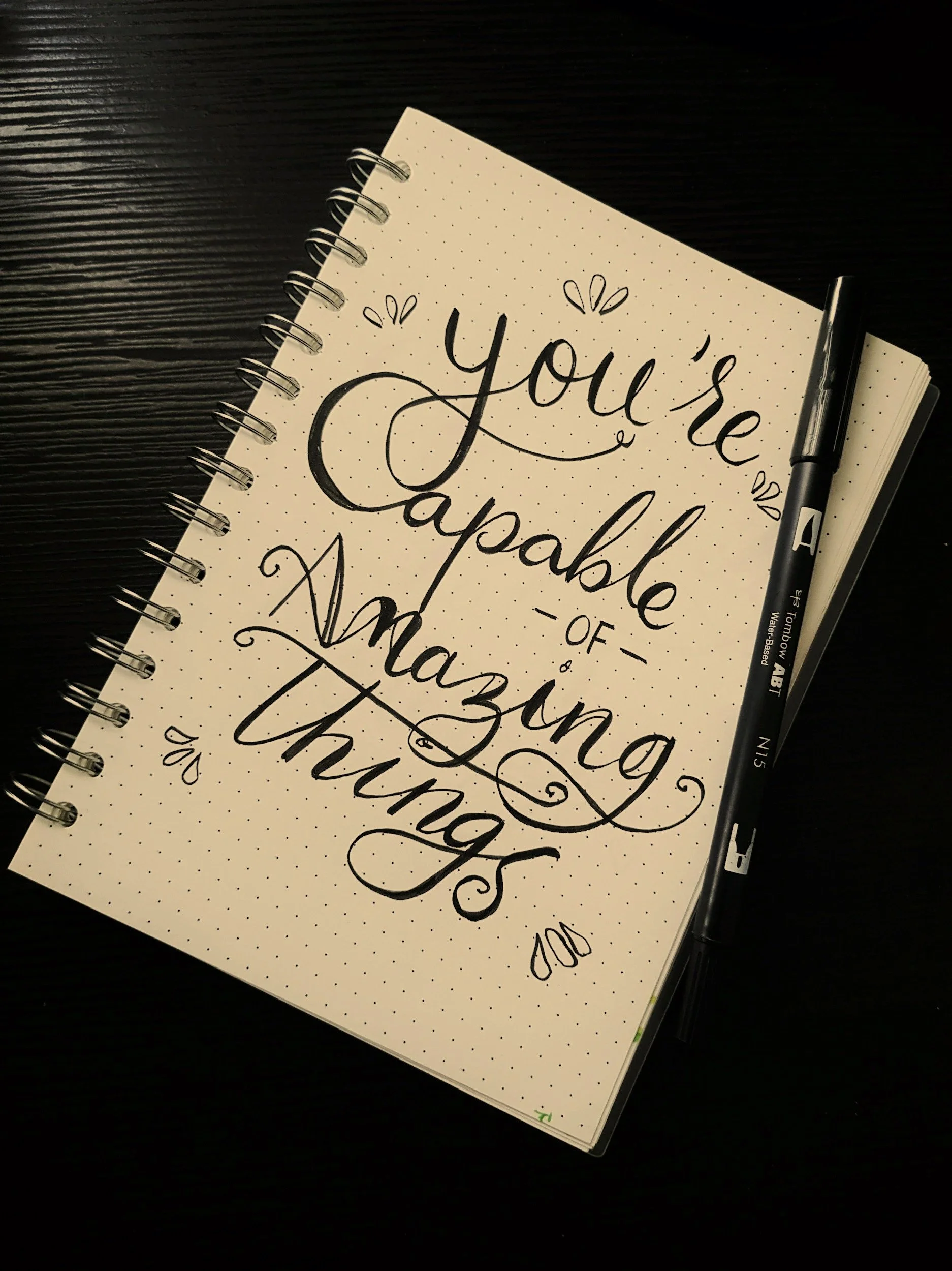 A brown notebook on a black table that says You're capable of amazing things with a black pen
