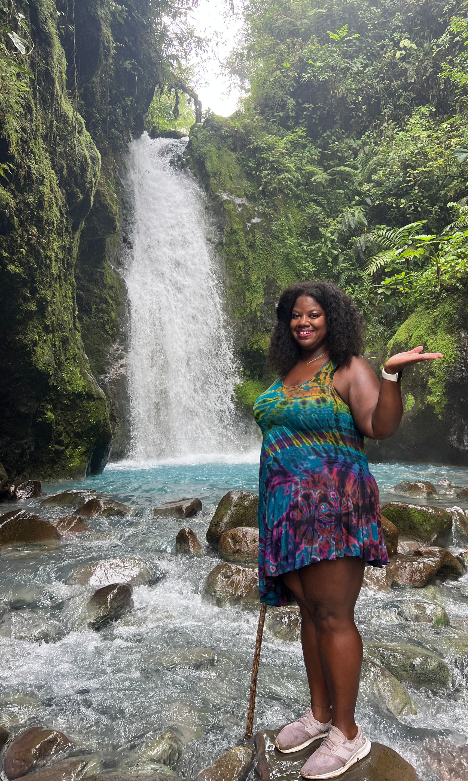 Lissette in a blue and purple tie dye dress standing in front of a waterfall with her left hand up and open