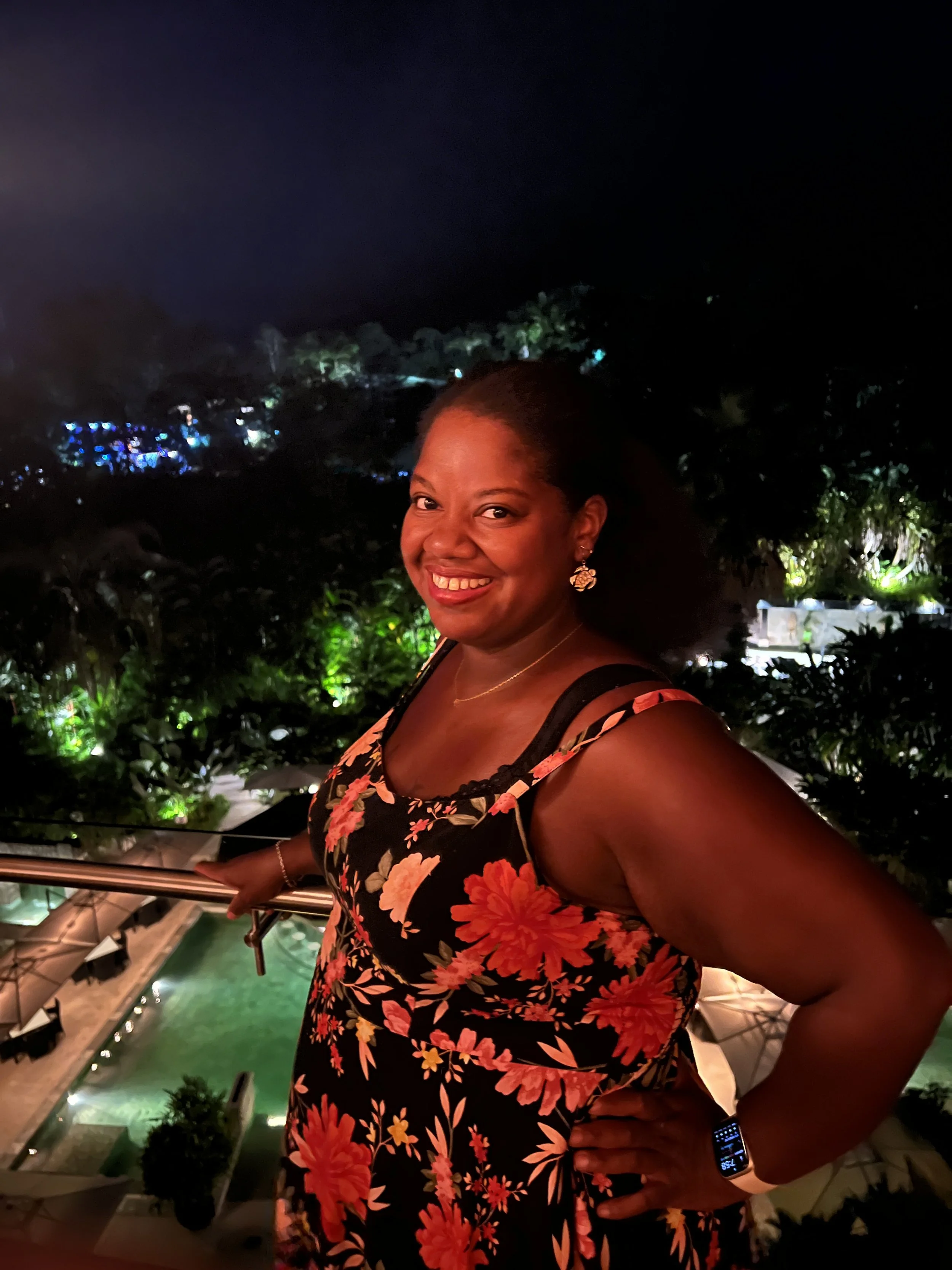Portrait of Lissette in a floral dressing holding a rail outside of a pool and trees at night
