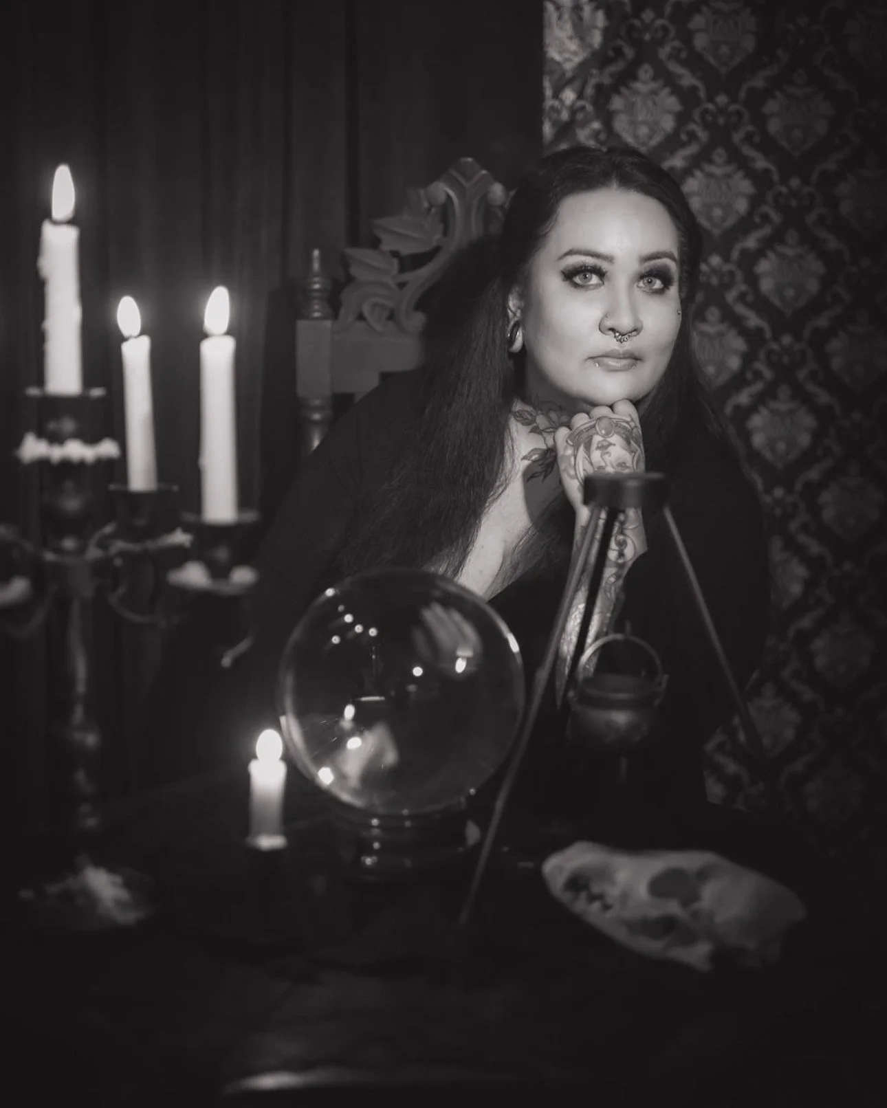 A woman with long dark hair, tattoos, and piercings, sitting at a table with candles, a crystal ball, and mystical objects, looking thoughtful in a dimly lit room with dark, ornate wallpaper.