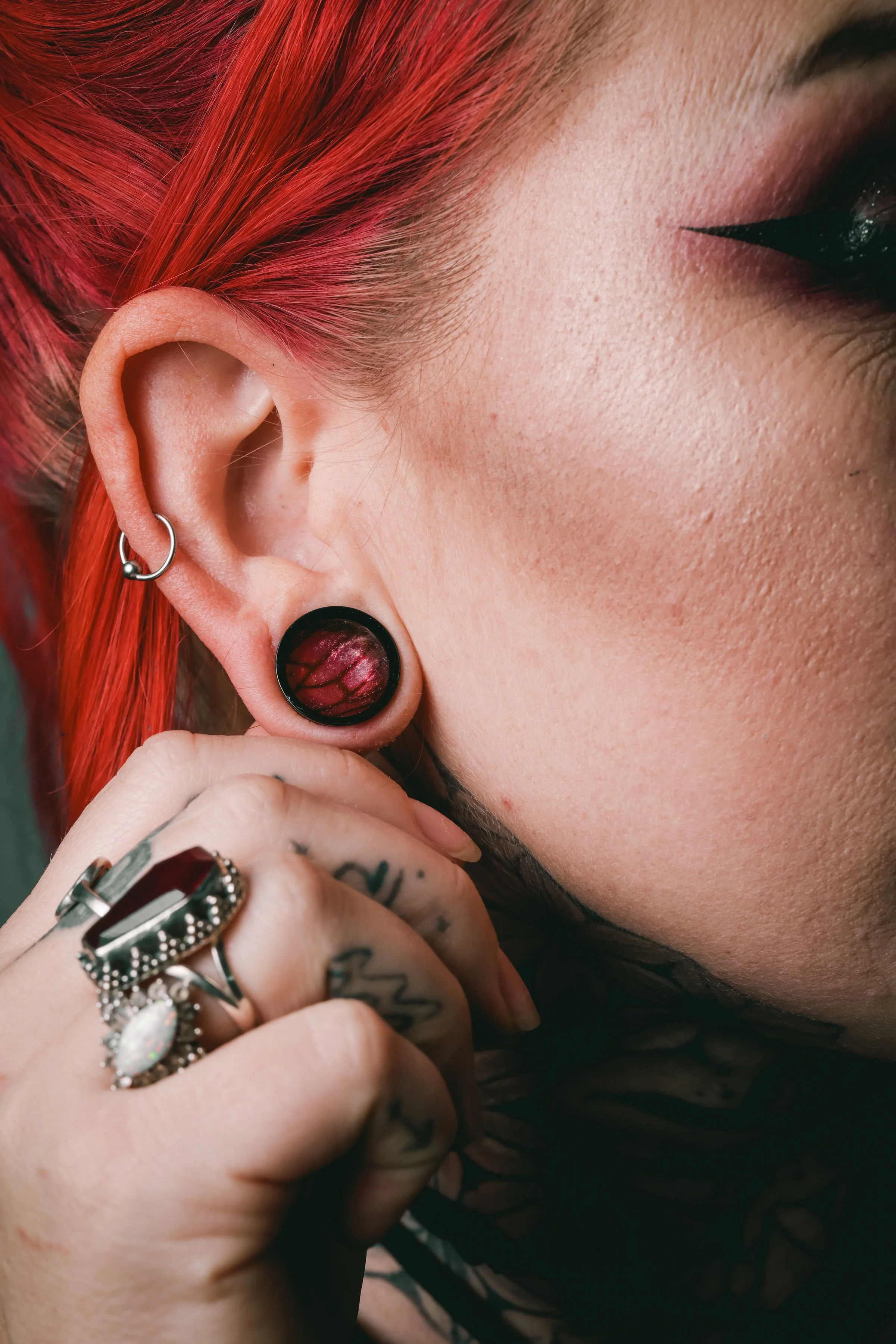 Close-up of a person's face showing their red hairstyle, black eyeliner, ear with a black plug earring and a hoop earring, and hand with multiple rings and tattooed fingers.