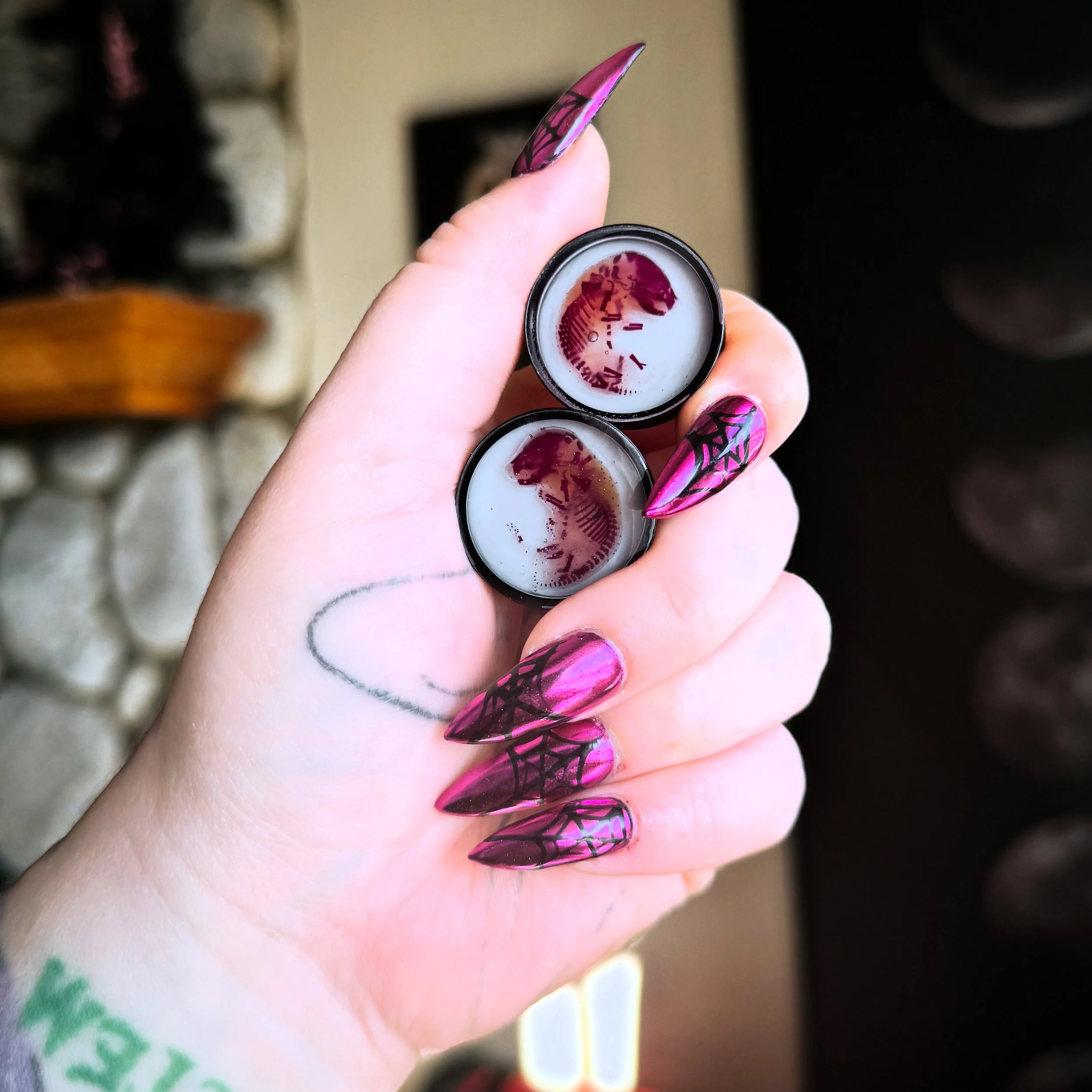 A hand with spider web themed nail art holding two small containers of white and red gel or paint, with a background of stone and wood.