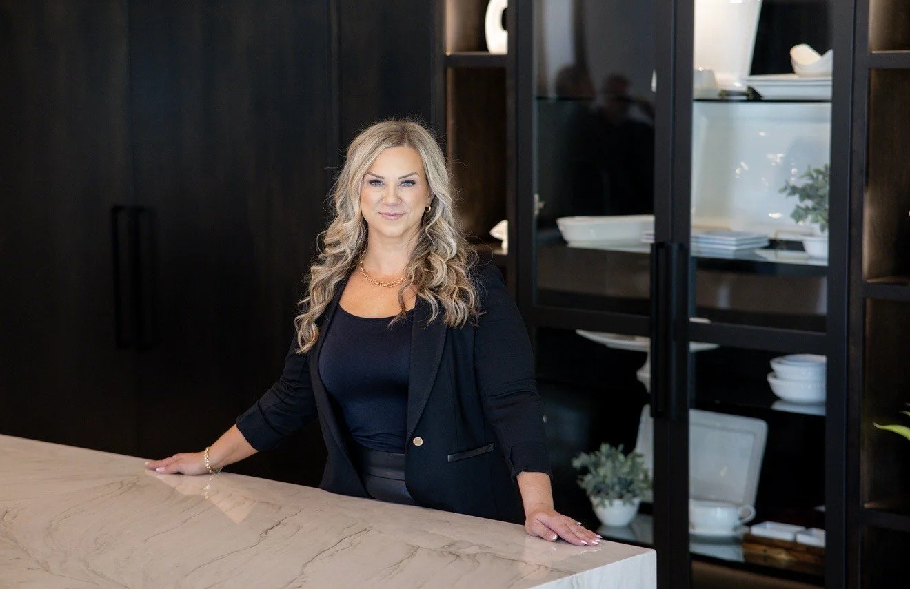 A professional woman with blonde hair in a black blazer standing behind a marble countertop in a modern kitchen or office space.