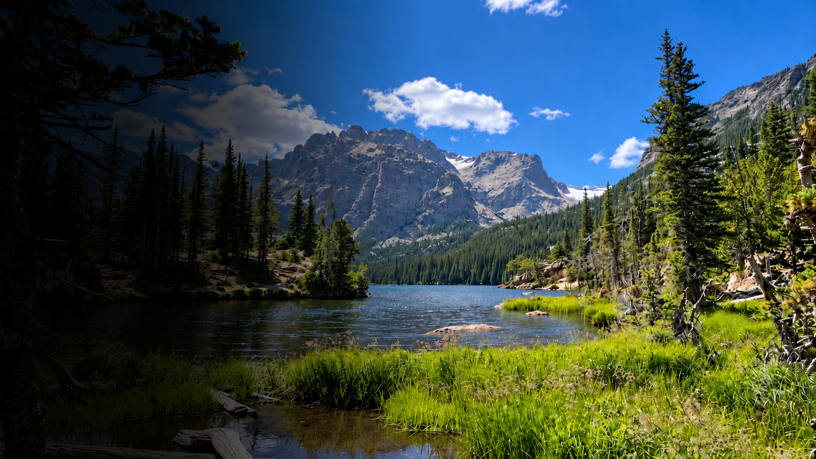A peaceful mountain lake scene with clear water, surrounded by lush green grass and tall pine trees. In the background, rugged mountain peaks rise against a blue sky with scattered clouds.