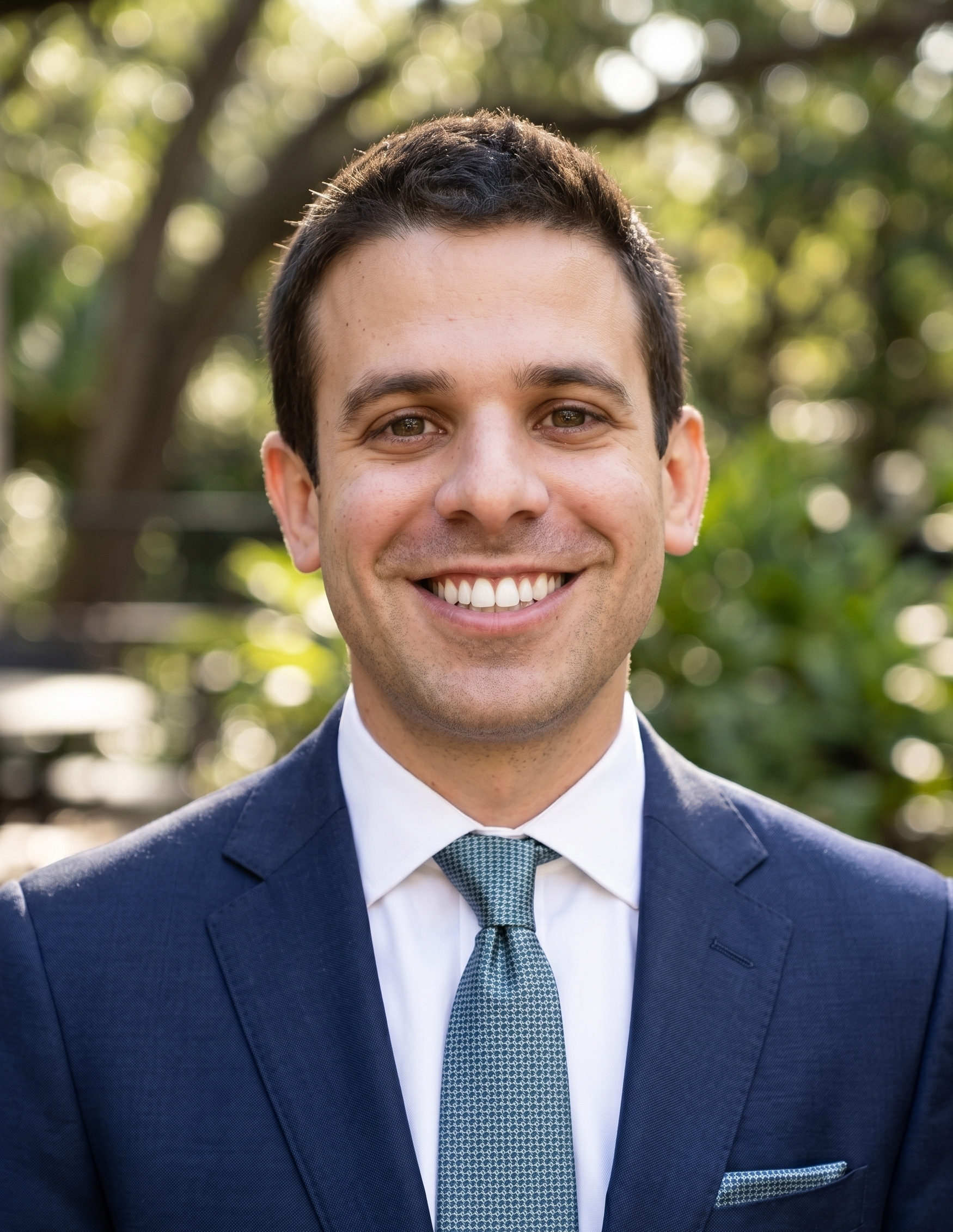 A young man in a navy blue suit smiling outdoors, with blurred trees and sunlight in the background.