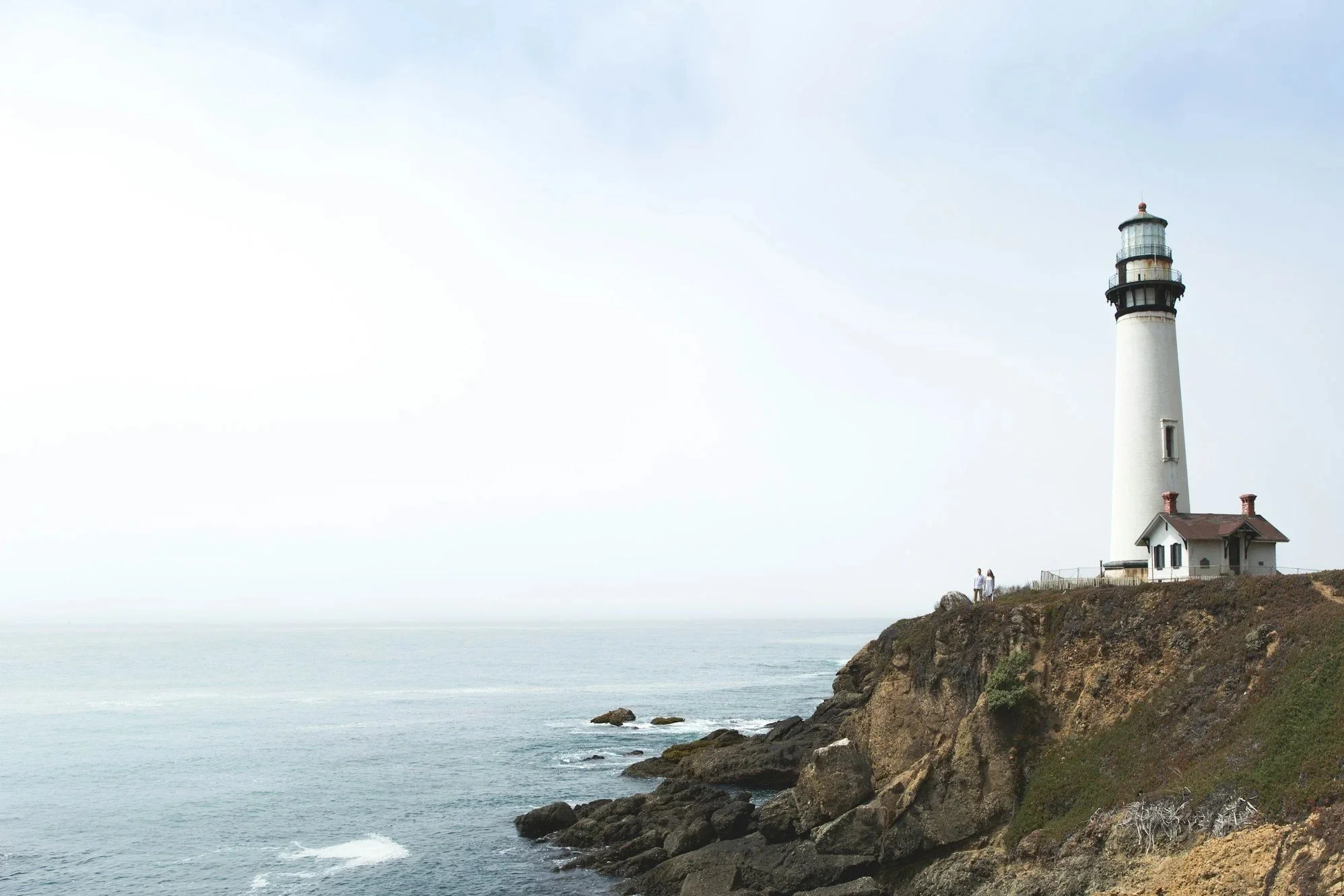 Lighthouse on a rocky cliff overlooking the ocean with two people walking nearby under a partly cloudy sky.