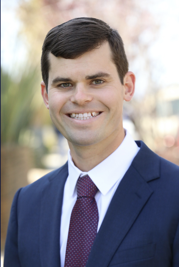 A man in a suit smiling outdoors with a blurred background of trees and sunlight.