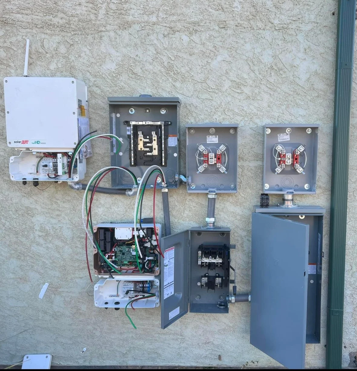 Electrical panels and wiring mounted on an exterior wall, some panels open revealing internal components and circuit boards.