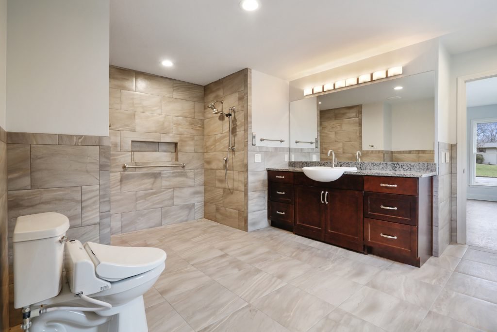 Bathroom with beige tile walls and floor, a walk-in shower area, a white toilet, a dark wood vanity with a granite countertop, a vessel sink, large mirror, and window showing outdoor greenery.
