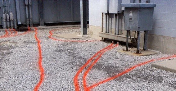 Gravel ground with orange spray-painted lines for utility markings near electrical boxes outside a building.