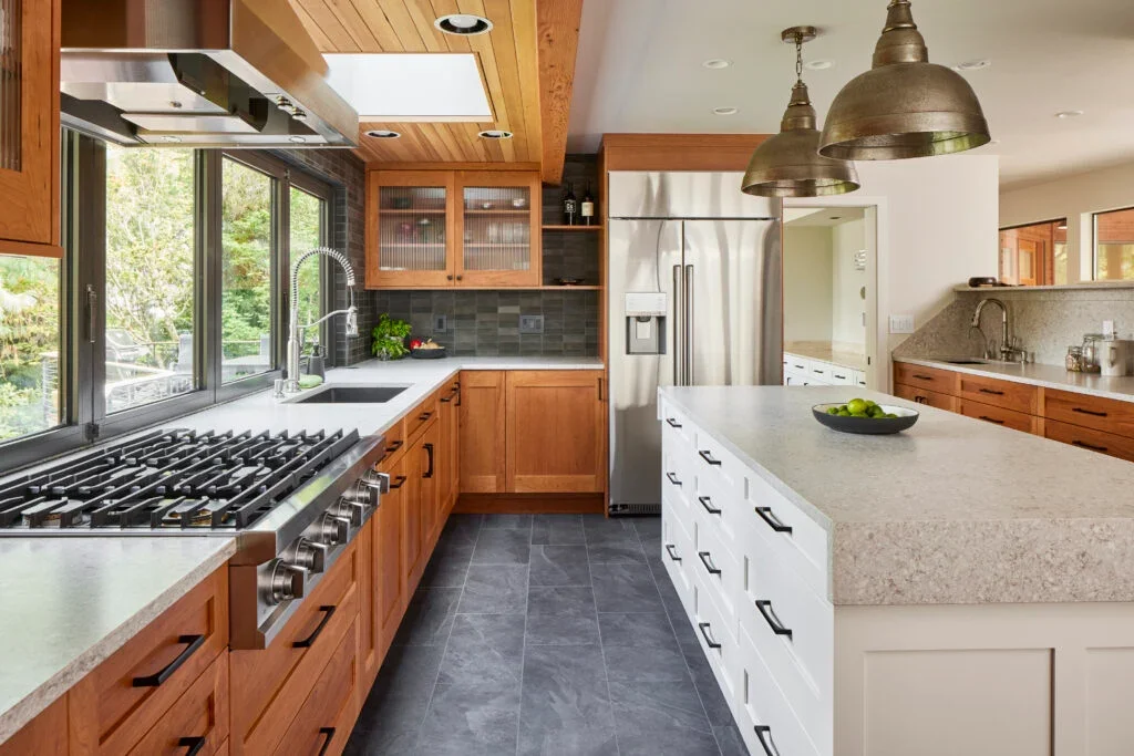 Modern kitchen with wooden cabinets, a large island with a light-colored countertop, stainless steel appliances, and large windows overlooking greenery.