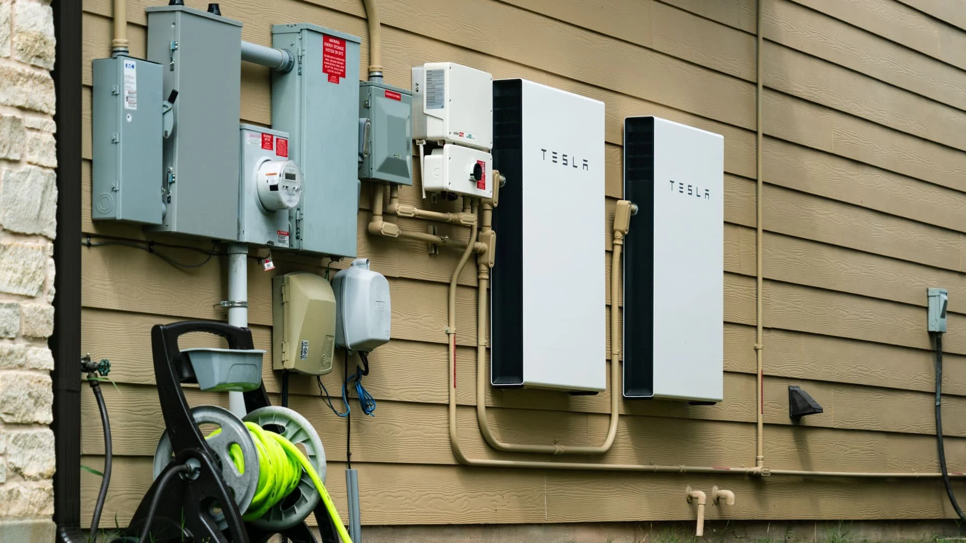 Electric meters, electrical panels, and Tesla power wall units installed on a house exterior wall, with a hose reel and garden hose on the ground.