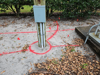 Electrical box on a pole with red spray paint markings on the ground around it, including a semicircular line, straight lines extending outward, and a circle at the base.