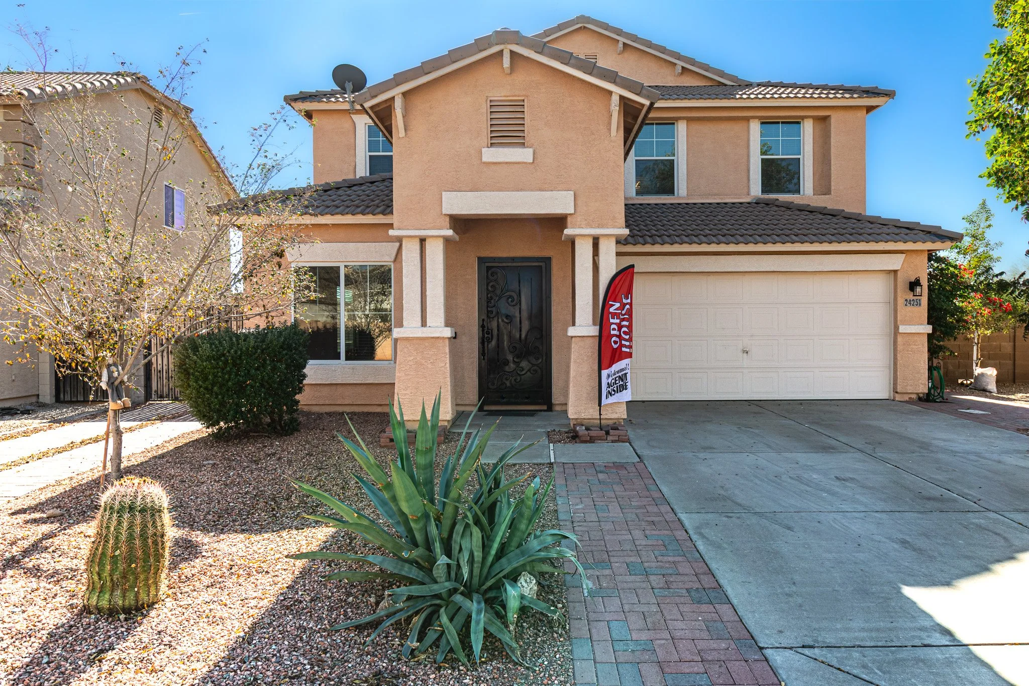 Front view of a two-story beige house with a large garage. A sign indicates the house is for sale, and the front yard features desert landscaping with a cactus, agave plant, small tree, and shrub. A brick pathway leads to the front door.