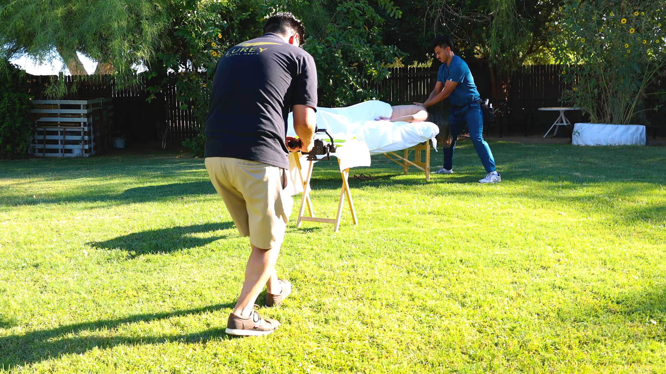 Two people performing CPR on a person lying on a table outdoors in a backyard with green grass and trees.