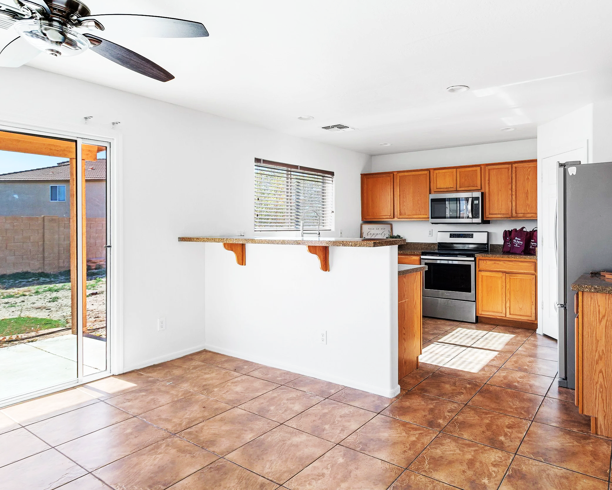 A bright kitchen with white walls, wooden cabinets, a stainless steel microwave, and a sliding glass door leading outside. There is a ceiling fan and tile flooring.