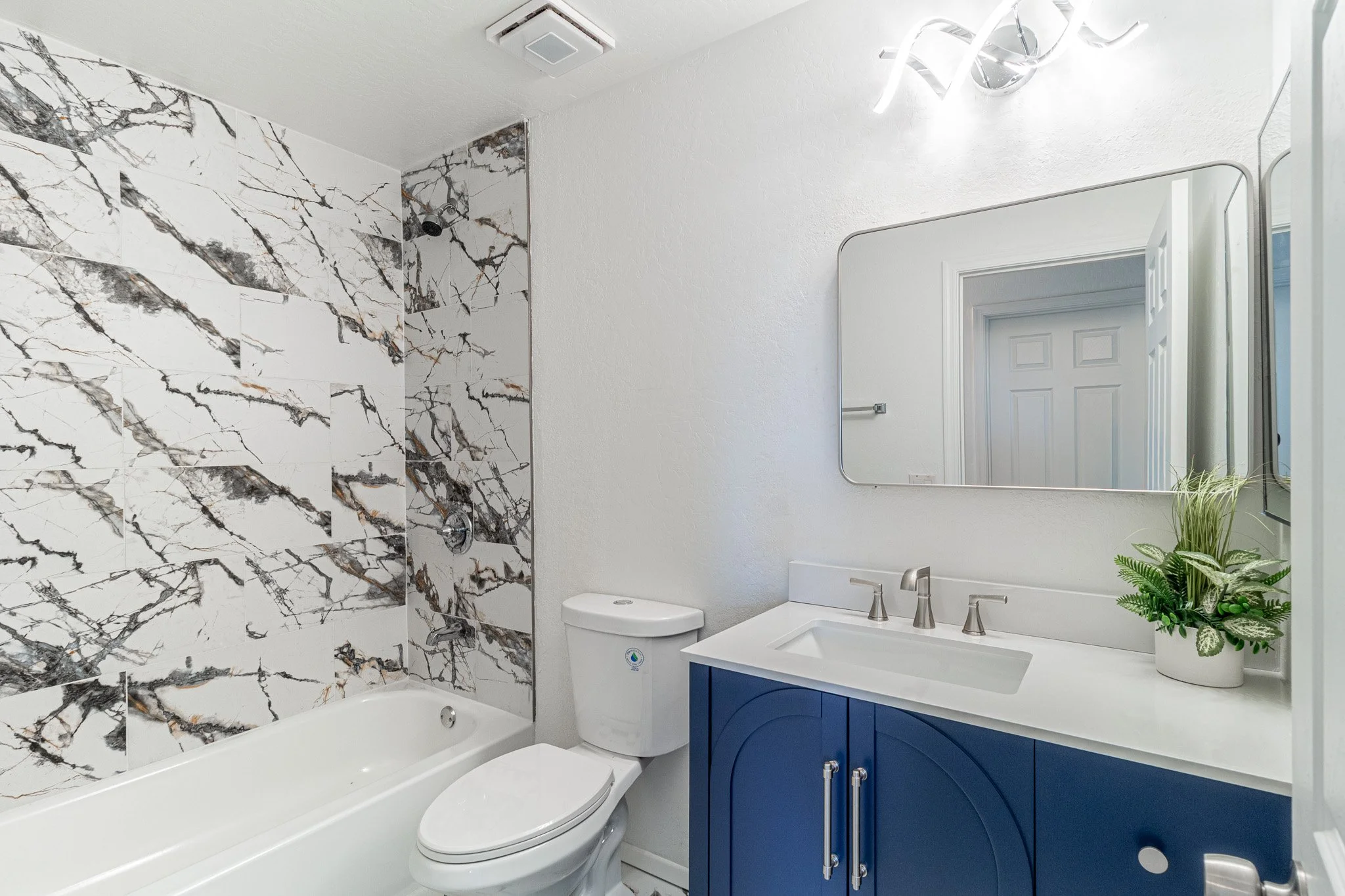 Modern bathroom with marble tiled shower, white toilet, navy blue vanity with white countertop, large mirror, and decorative green plants.