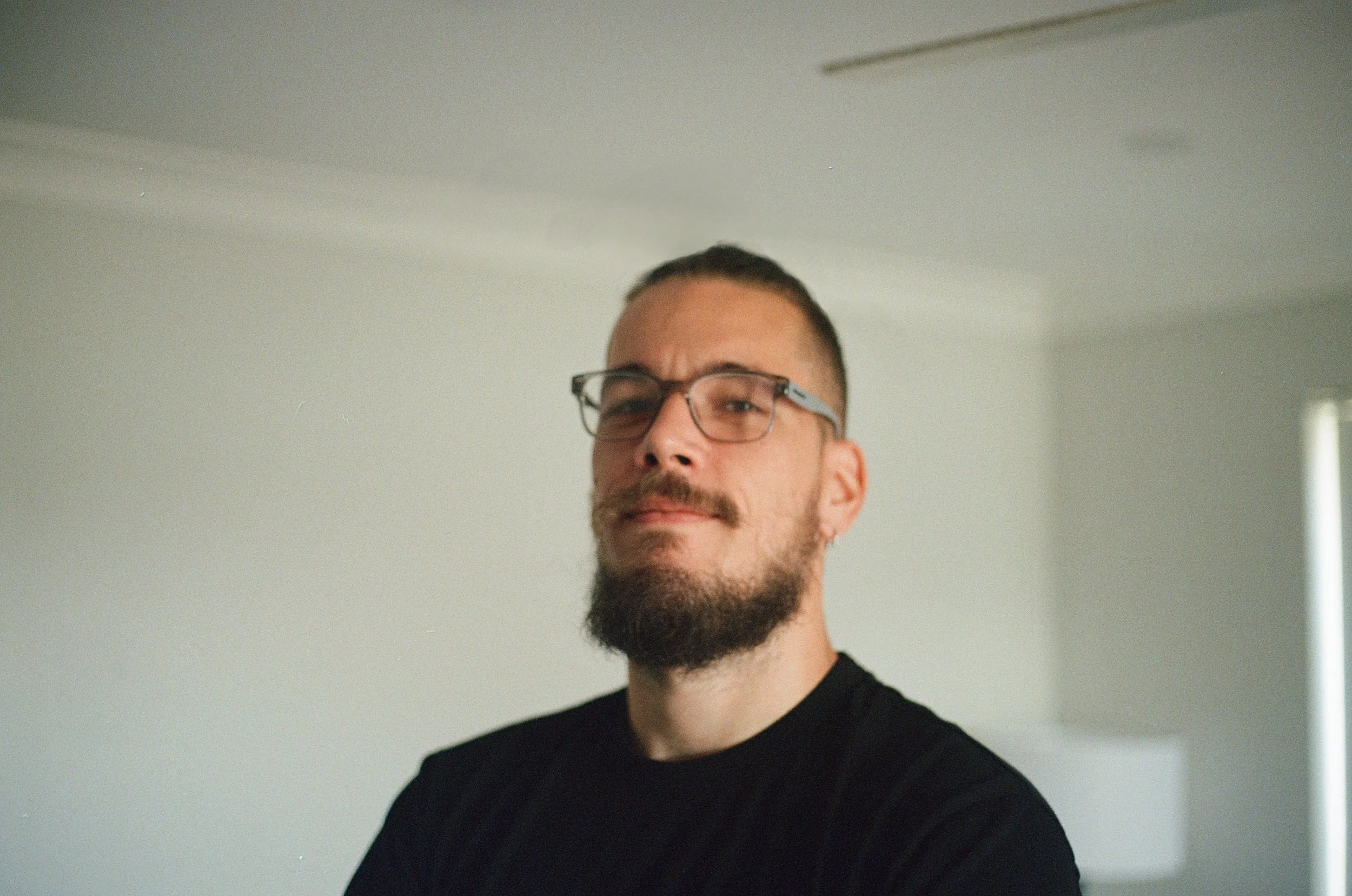 Portrait of a man with glasses, beard, and mustache, wearing a black shirt, standing in a room with neutral walls and a window.