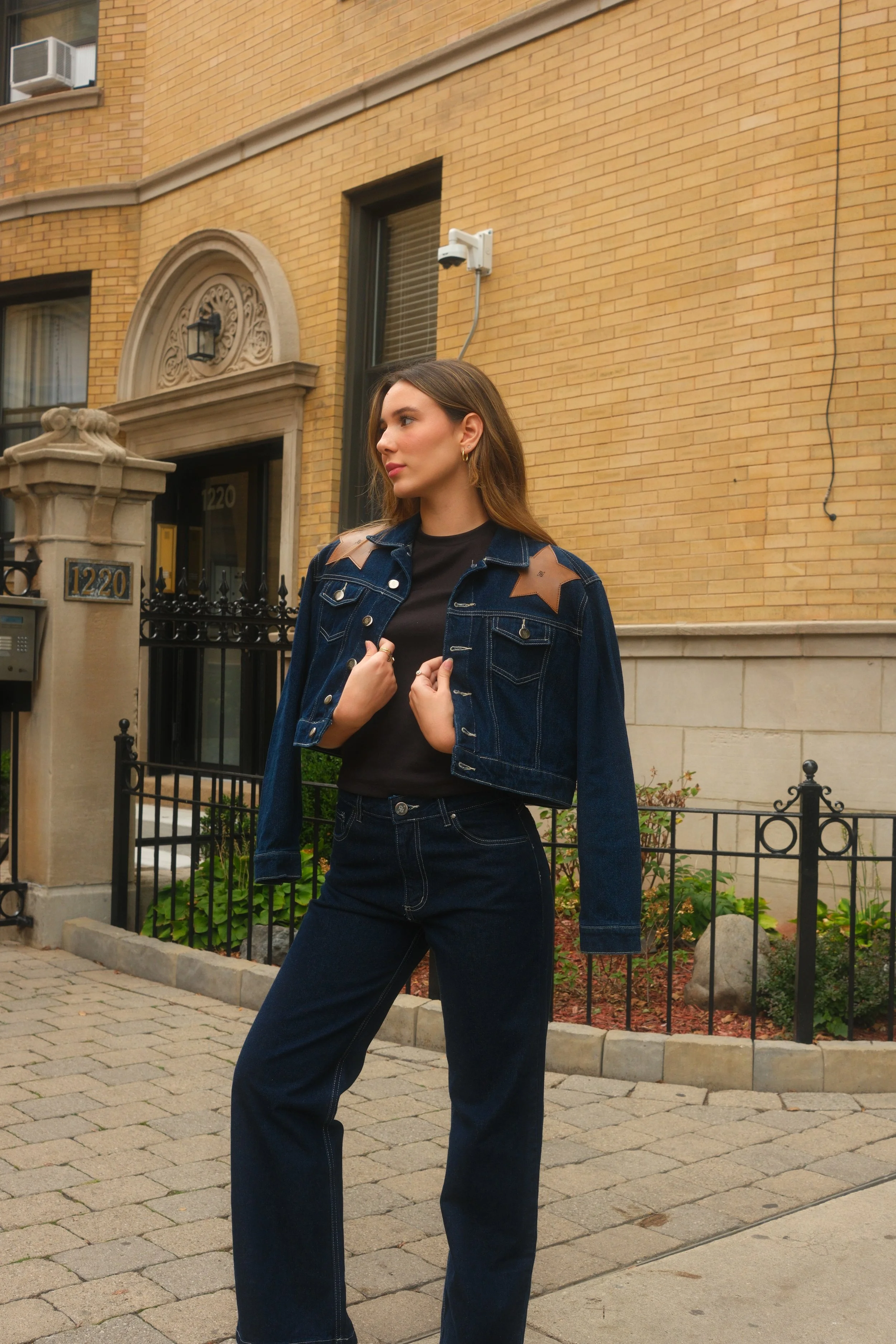 Young woman in dark blue denim jacket with shoulder patches and matching jeans, standing on a city sidewalk near a brick building with window air conditioner units.