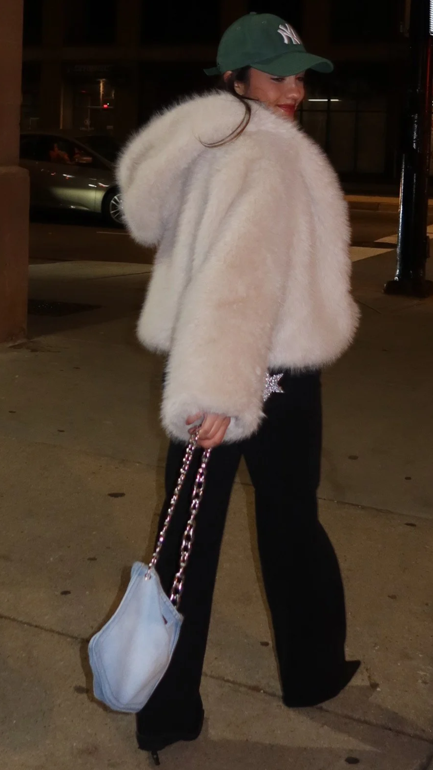 Woman in a white fluffy fur coat, black pants, green baseball cap with a white logo, holding a white purse with a chain strap, standing on a sidewalk at night.