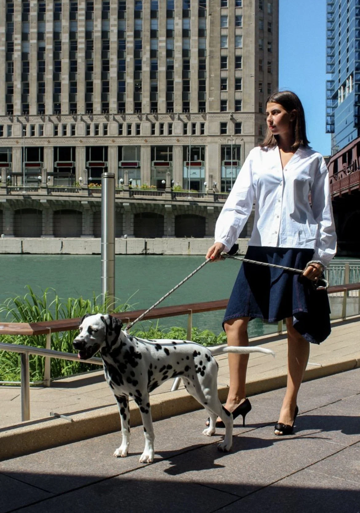 A woman walking a Dalmatian dog along a city sidewalk beside a river, with tall buildings in the background.