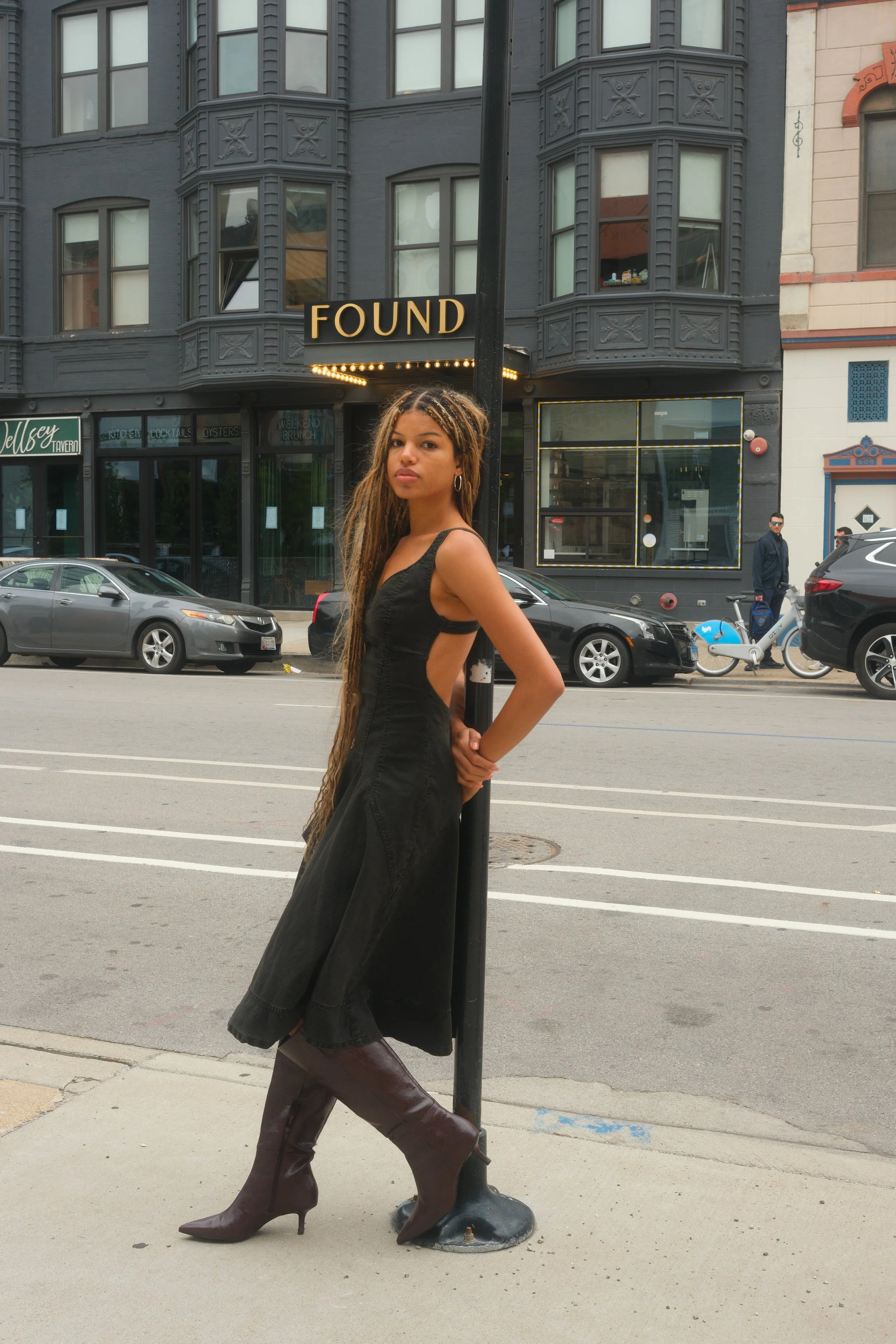 A young woman with long dreadlocks standing on the sidewalk, leaning against a lamp post with a 'FOUND' sign, in an urban setting with parked cars and buildings in the background.