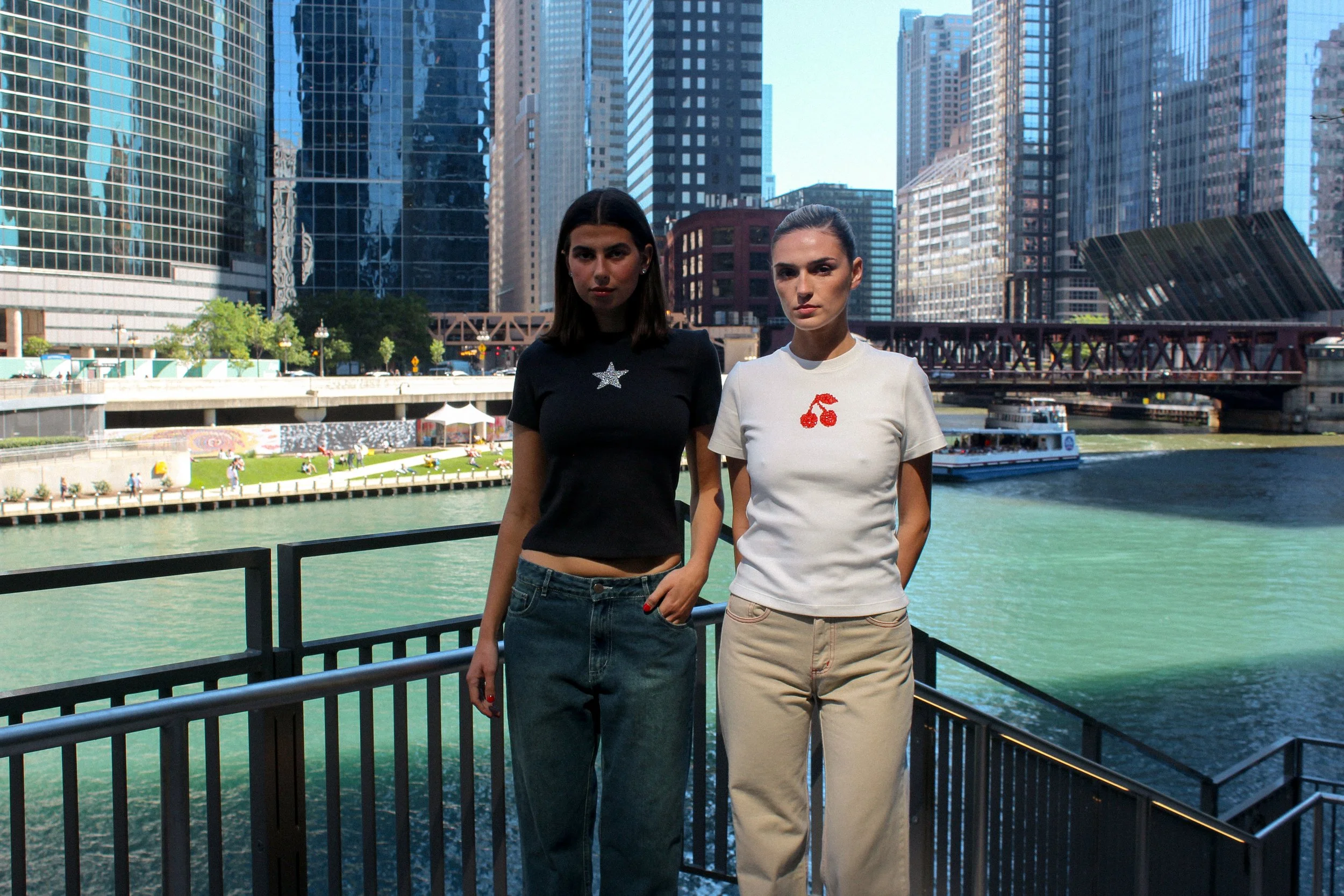 Two women standing by a railing near a river with city skyscrapers and a red bridge in the background