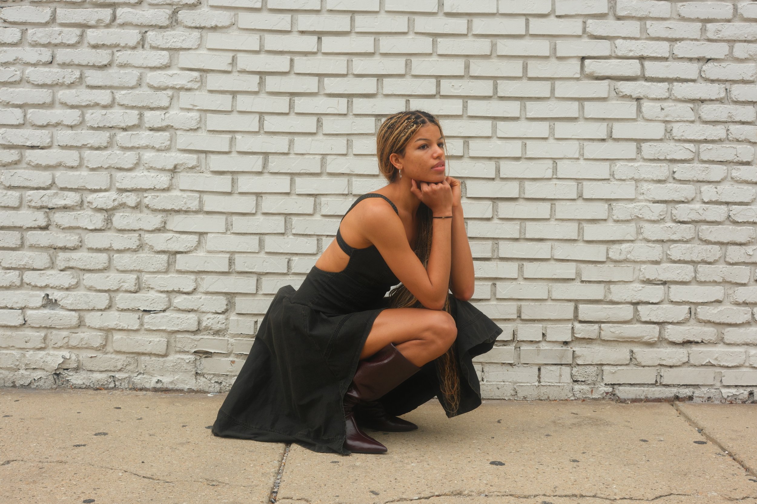 A woman squatting in front of a white brick wall, wearing a black dress and boots, with her chin resting on her hands, looking thoughtfully into the distance.