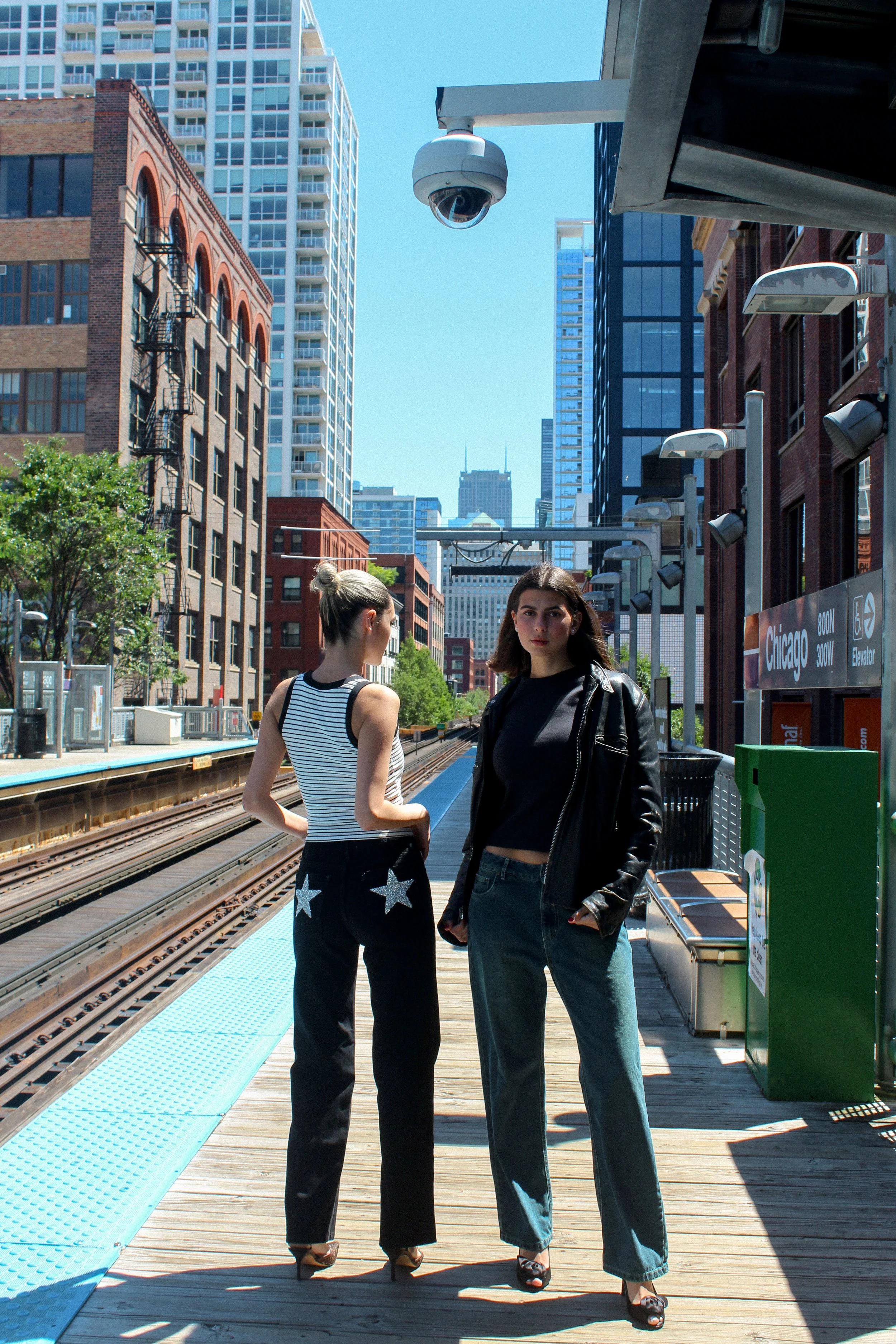 Two women standing on a Chicago train station platform with city high-rise buildings in the background, one in a striped tank top and star-embellished pants, the other in a black leather jacket and jeans, under a clear blue sky.