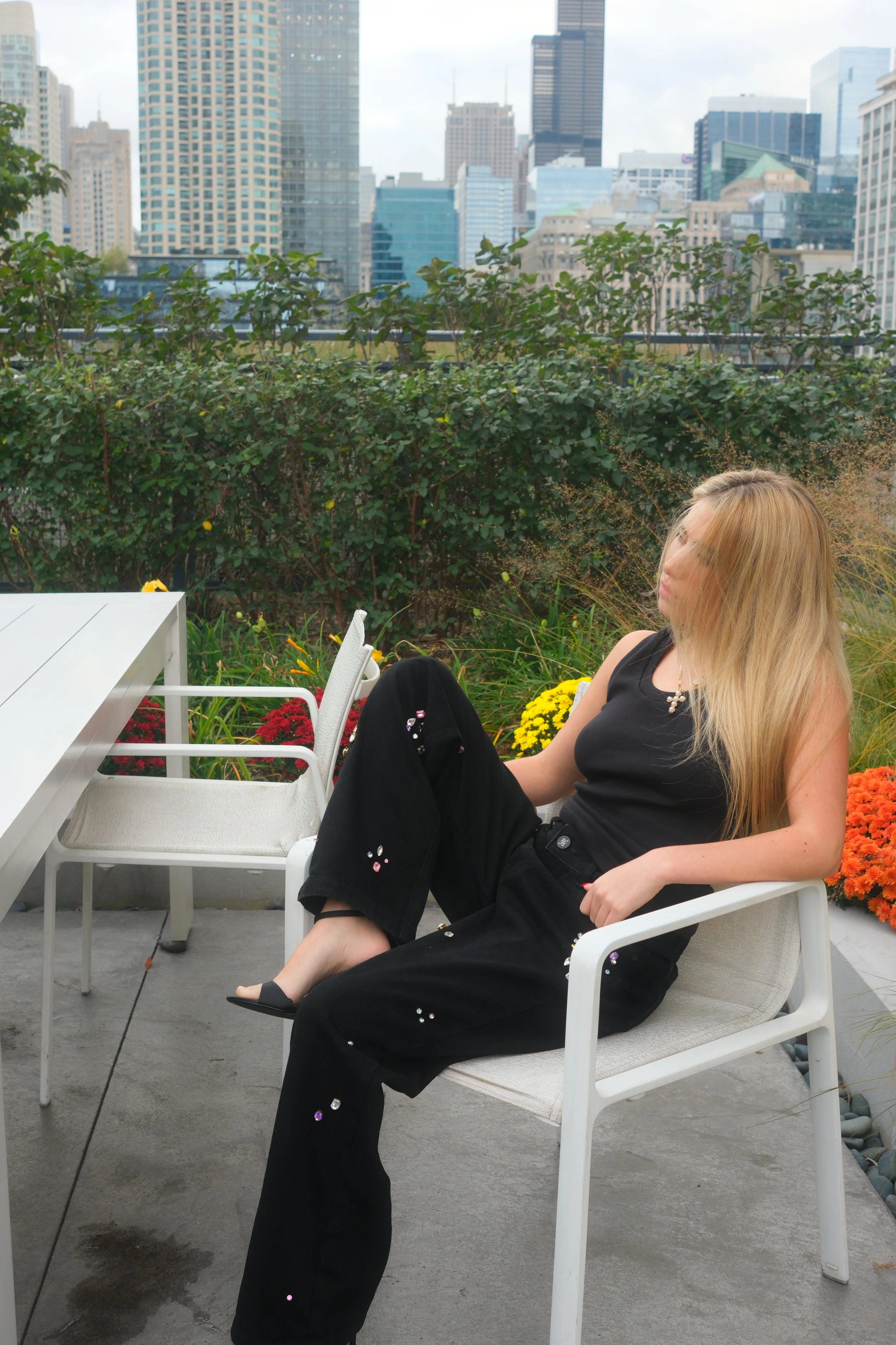 A woman with long blonde hair sitting on a patio chair in an urban rooftop garden, with a city skyline in the background, plants, and flowers around her.