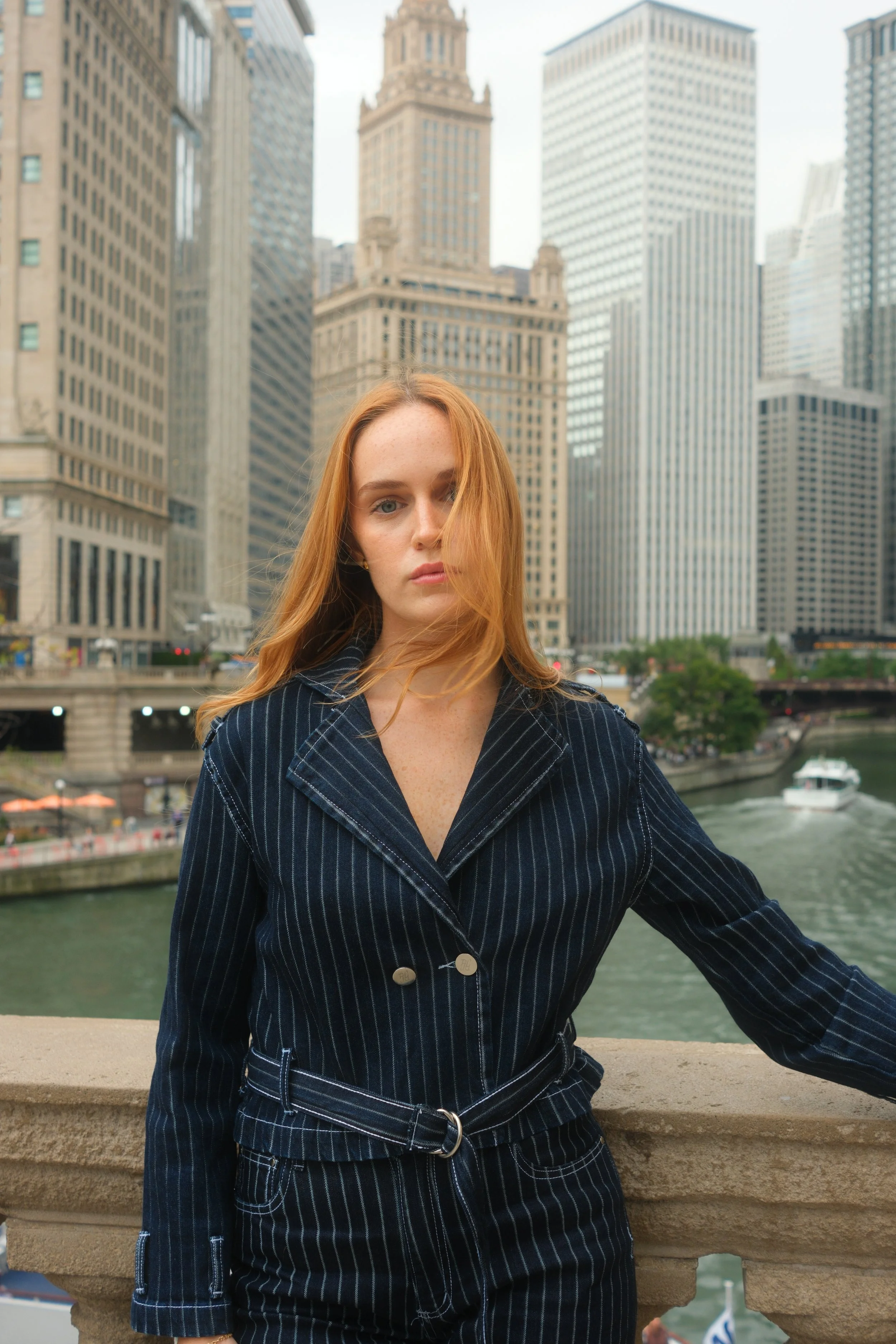 A young woman with red hair and freckles, wearing a dark pinstripe suit, stands on a city bridge with skyscrapers and a river in the background.