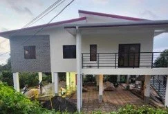 Two-story house with white walls and dark balcony railings, elevated on stilts, with a porch area and lush greenery around.