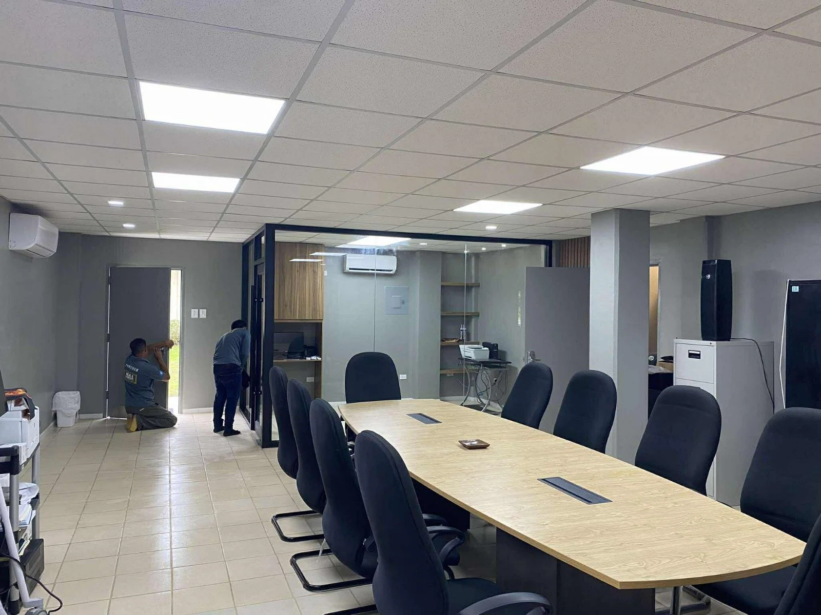 Two men working inside an office, one talking on the phone while kneeling and the other inspecting a wall or partition.