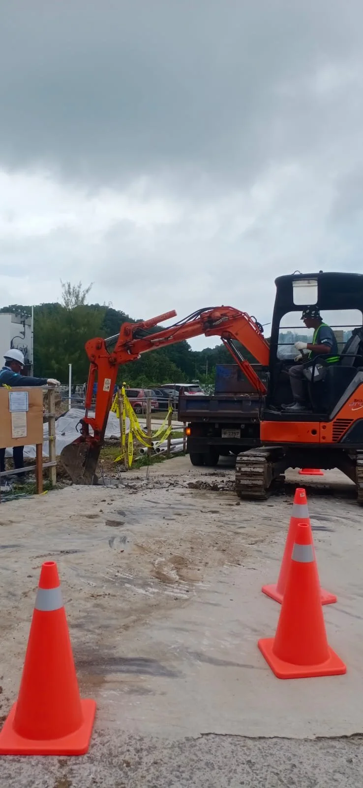 Construction workers operating an orange mini excavator at a construction site, with obstacles and safety cones visible, under cloudy skies.