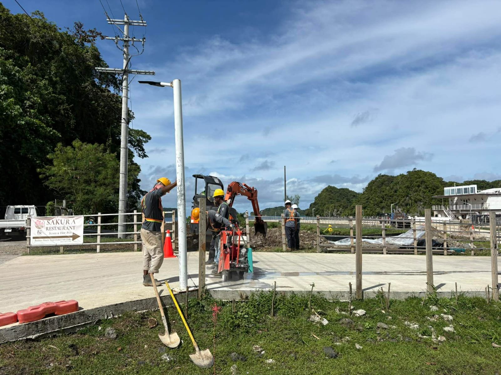 Construction workers installing or repairing utility poles on a sidewalk with construction tools and equipment around, nearby a fenced construction site under a partly cloudy sky.