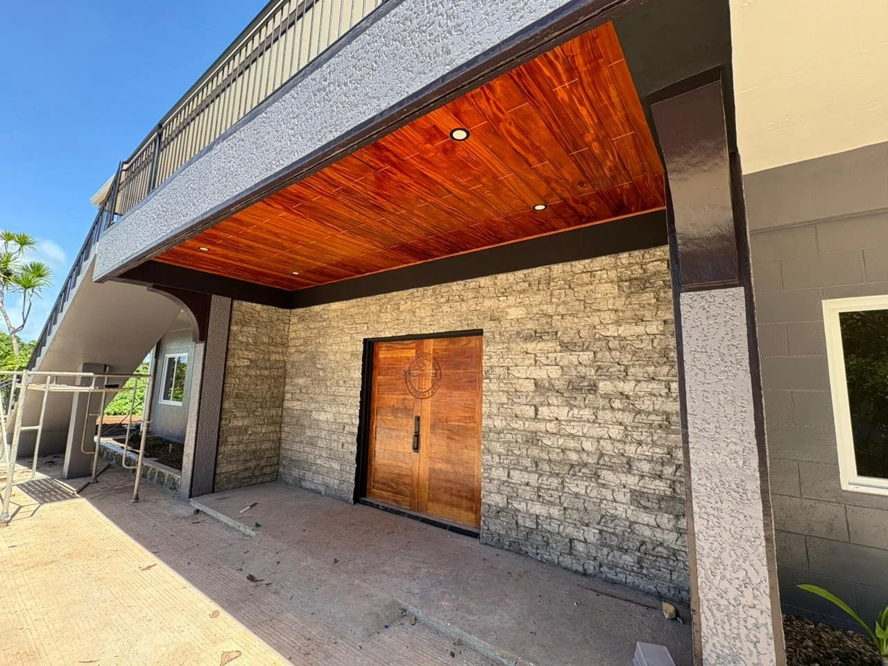 Modern building entrance with stone wall, wooden double doors, and a curved balcony above with a wood ceiling and black railing under a clear blue sky.