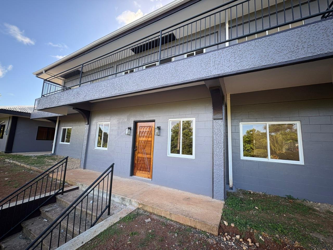 Modern gray two-story house with black railings and a wooden front door, surrounded by small patches of grass and a concrete staircase leading to the entrance, under a blue sky with clouds.
