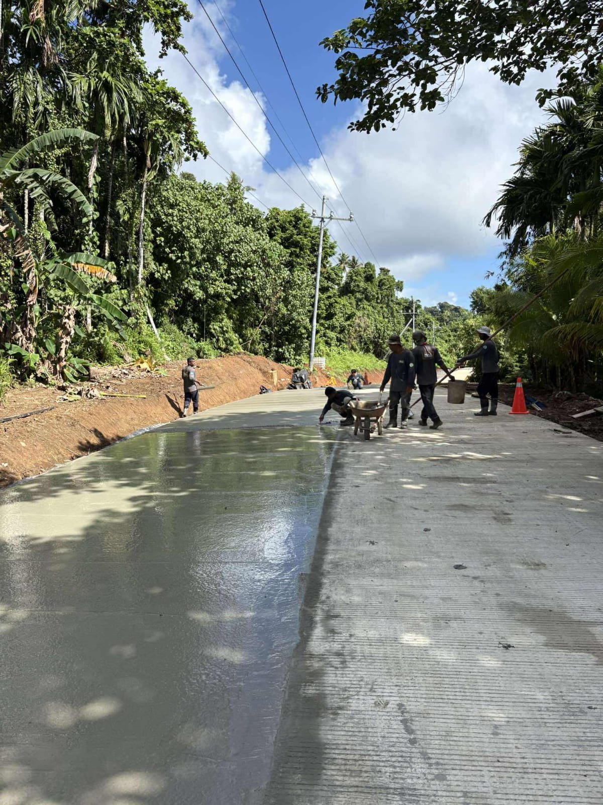 Construction workers are applying a layer of concrete on a rural road surrounded by lush green trees, with utility poles and wires overhead, under a partly cloudy sky.