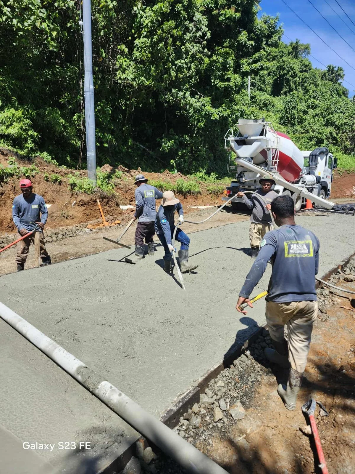 Workers pouring and leveling concrete on a road construction site surrounded by green trees.