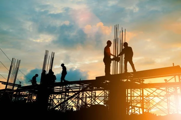 Construction workers silhouetted against a sunset sky working on a building's steel framework.
