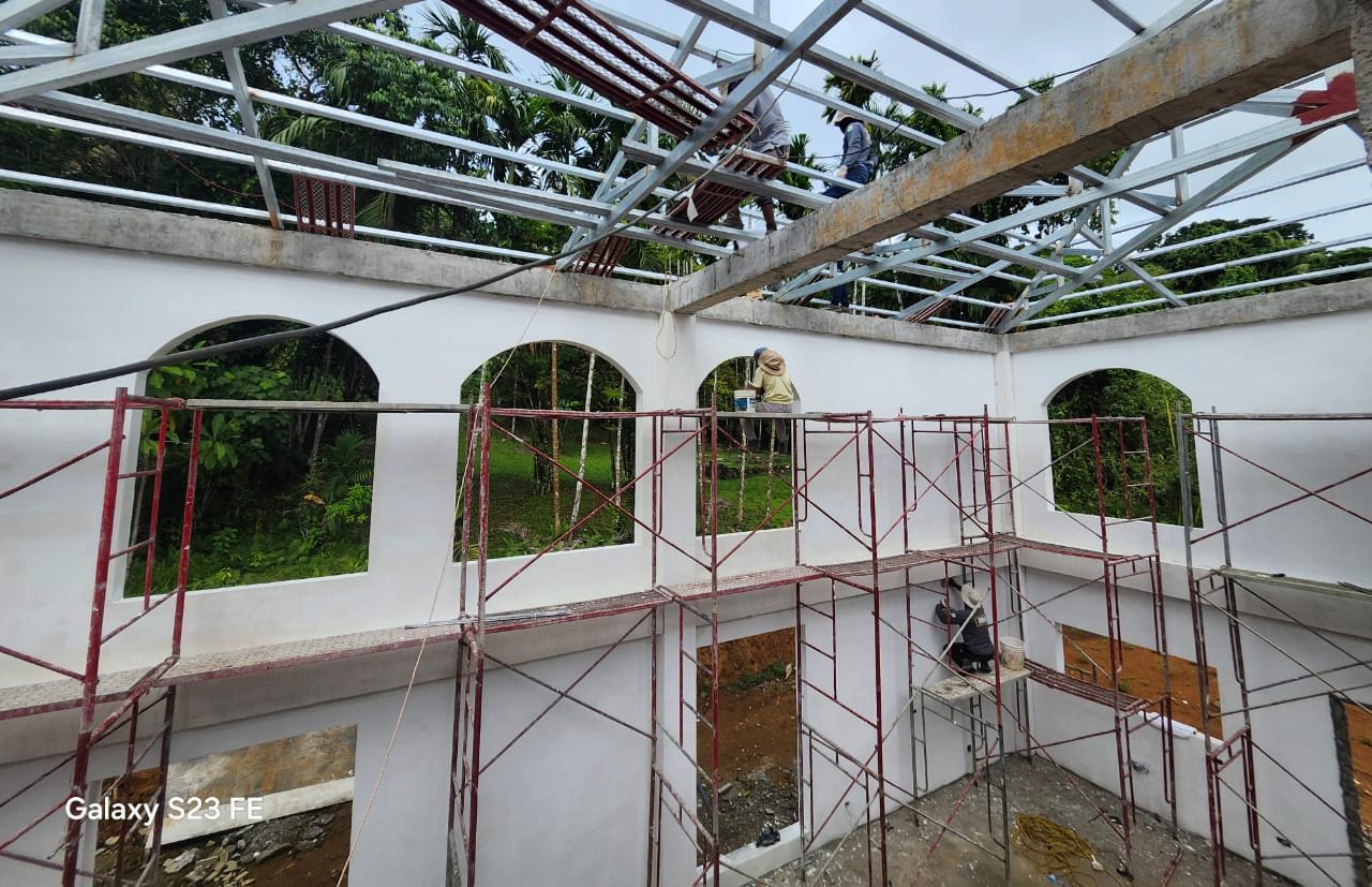 Construction site of a building with scaffolding and workers installing a metal roof framework, surrounded by greenery.