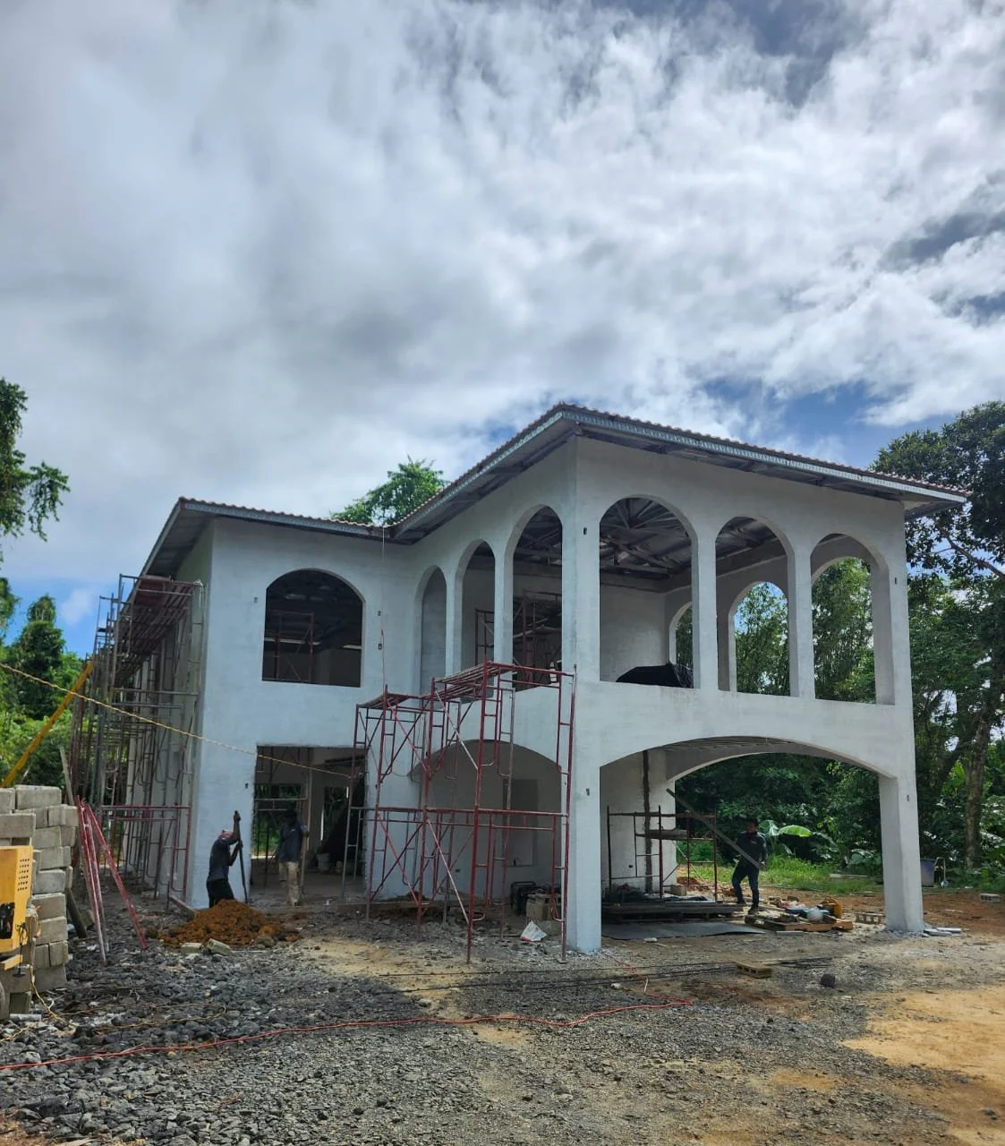 Construction of a two-story house with white walls, large arched windows, and red scaffolding around the building, with workers present on site and a cloudy sky overhead.