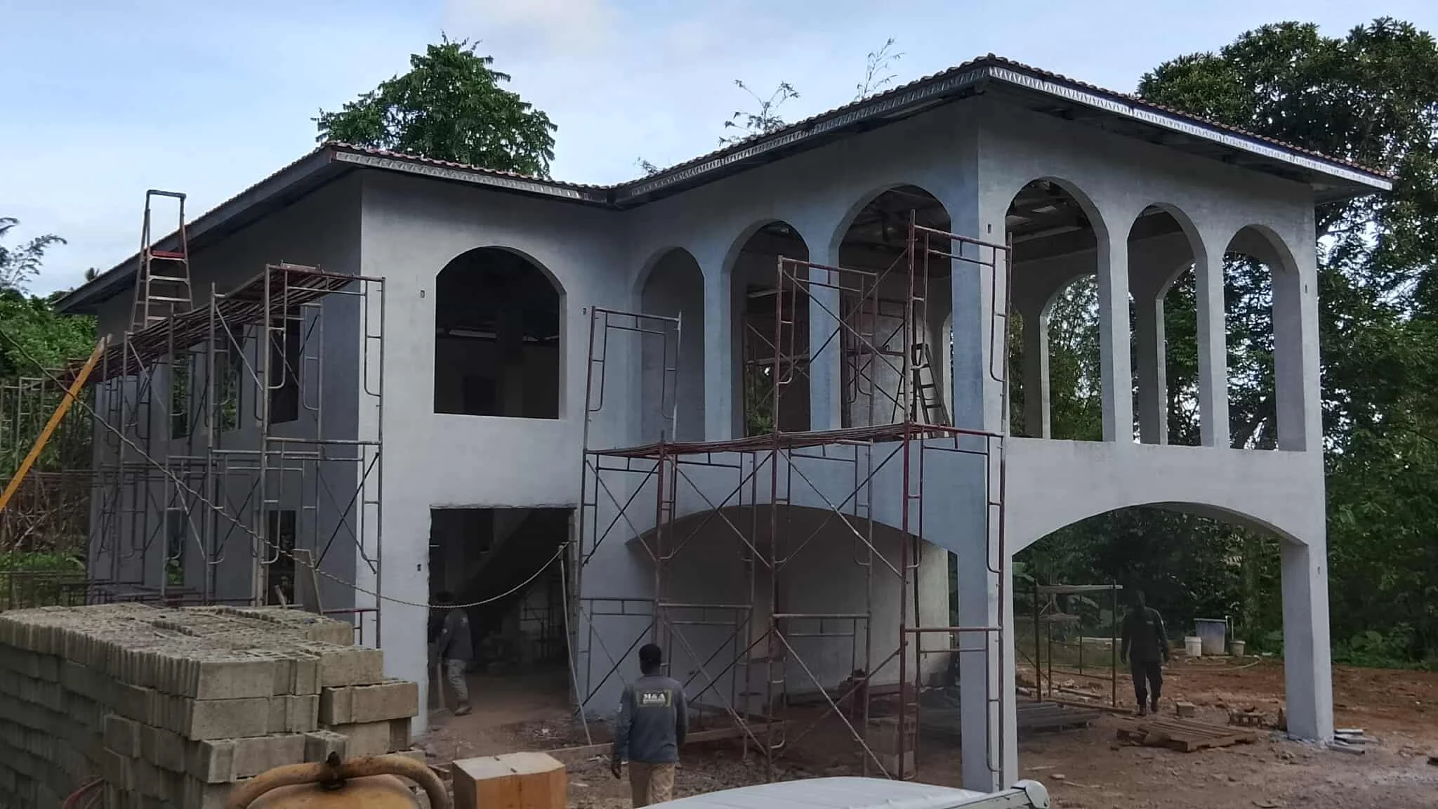 Under construction two-story house with scaffolding around it, arched windows, and a tiled roof, surrounded by trees and construction materials.