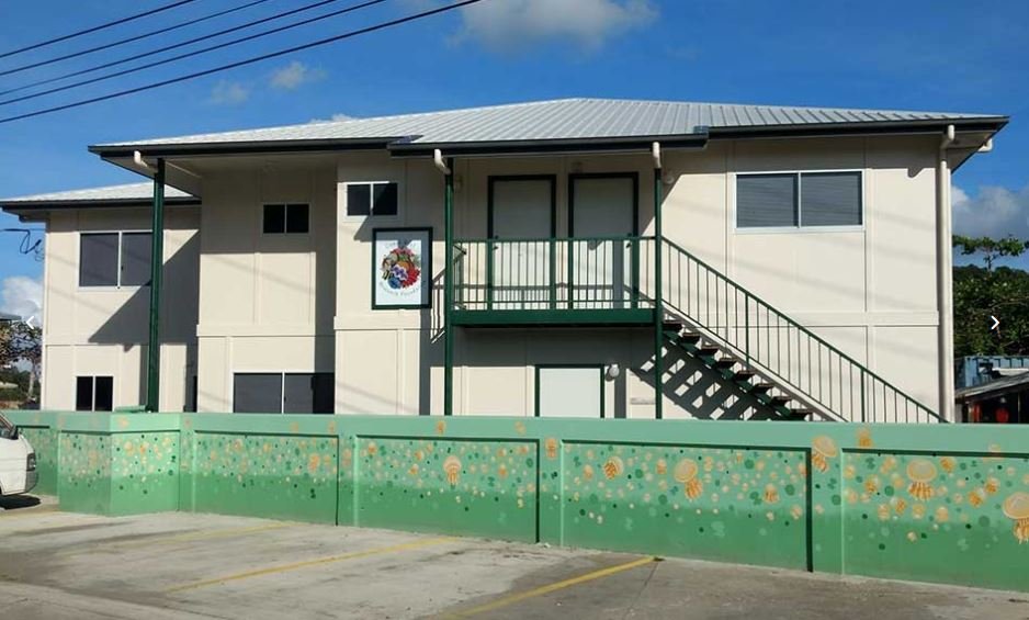 Two-story beige building with green stairs, decorated fence with jellyfish pattern, parking lot, and a colorful wreath on the wall.