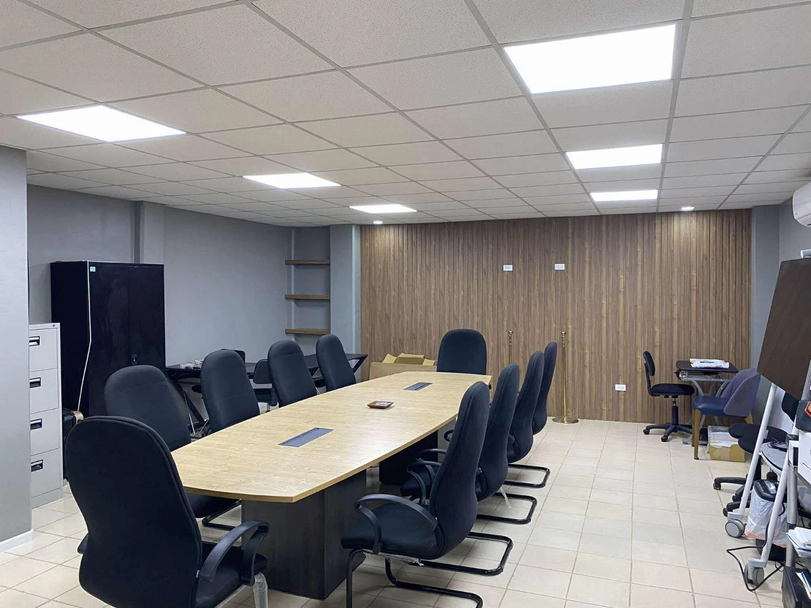 Empty conference room with a large wooden table, black office chairs, a gray wall, and a wooden accent wall at the back.