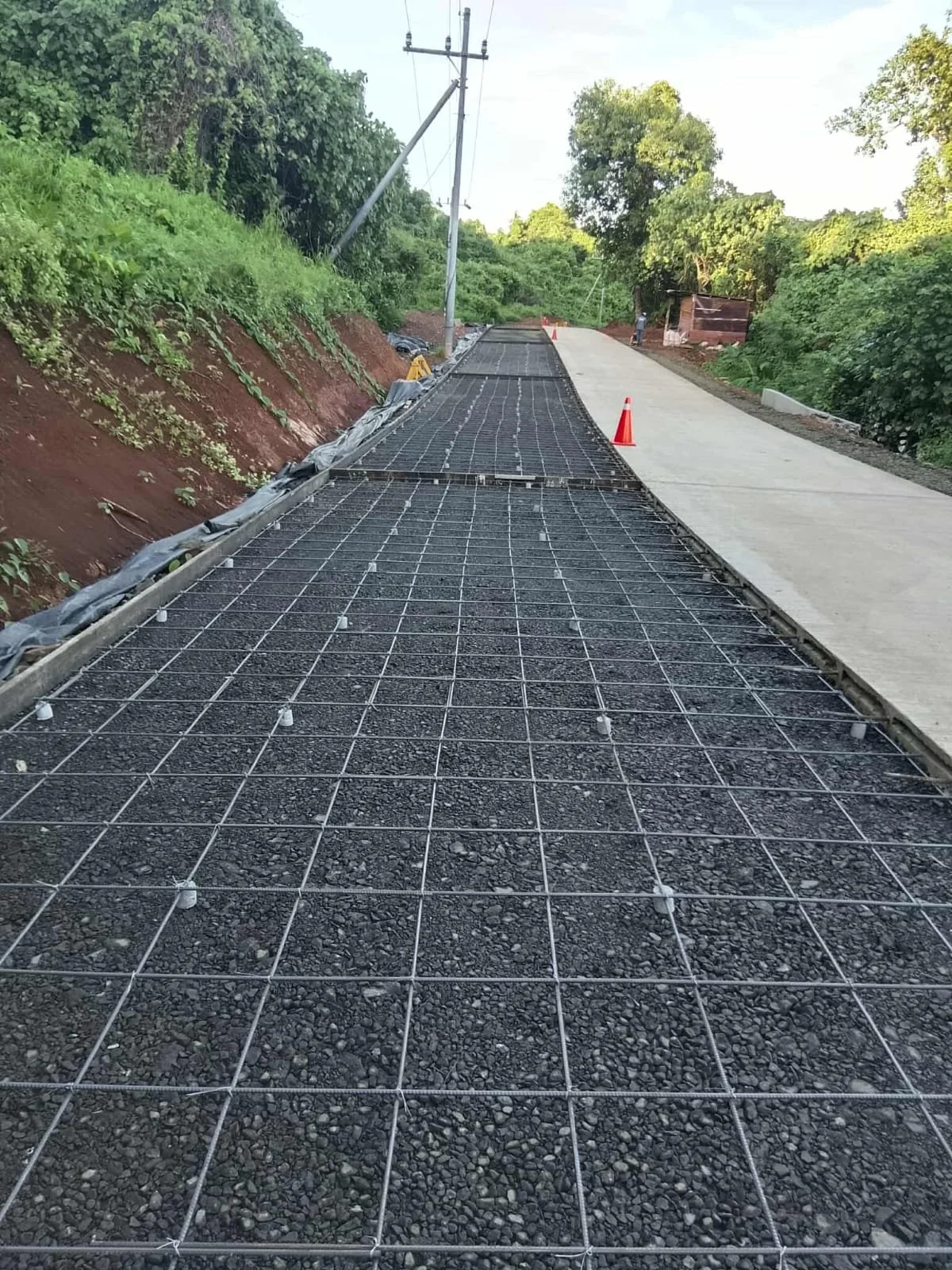 Construction site with newly installed concrete sidewalk and reinforced gravel surface, orange traffic cones, green trees, power lines, and blue sky.