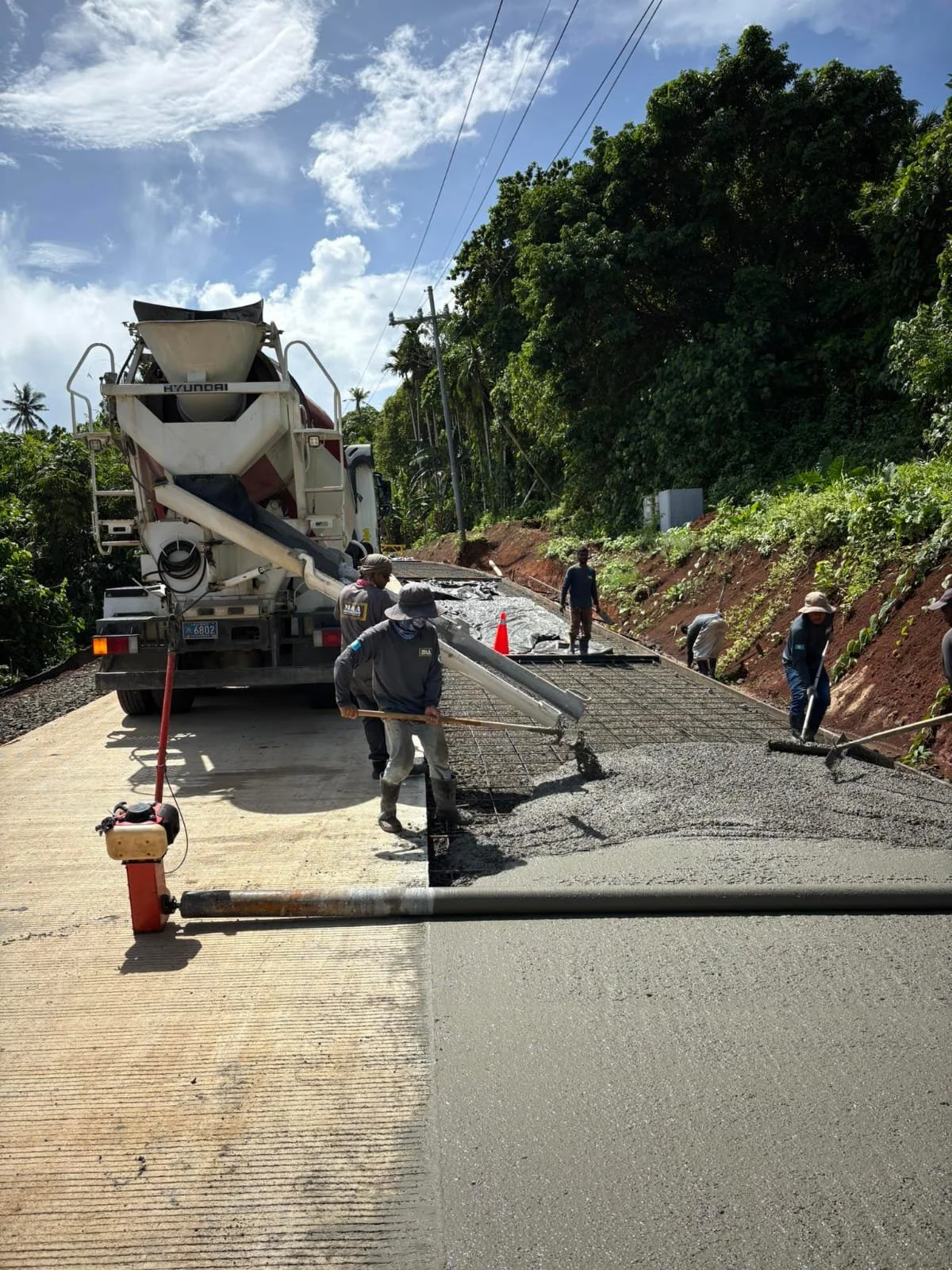 Workers pouring concrete on a road with a concrete mixer truck nearby, surrounded by green trees and a partially cloudy sky.