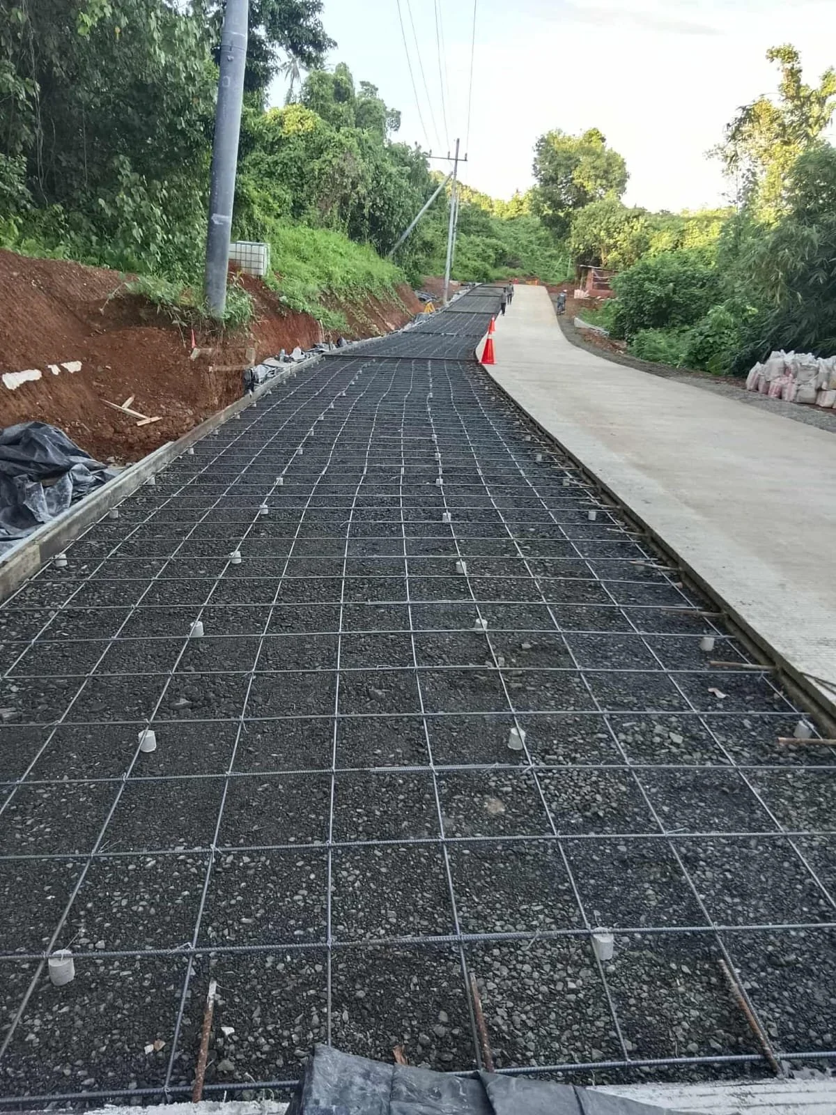 Construction site on a sloped road where concrete is being prepared for pouring, with a metal grid and plastic spacers, and a section of finished concrete pavement on the right.